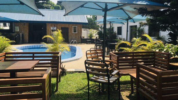Zona de piscina exterior con bancos de madera y sombrillas, rodeada de vegetación y edificios.