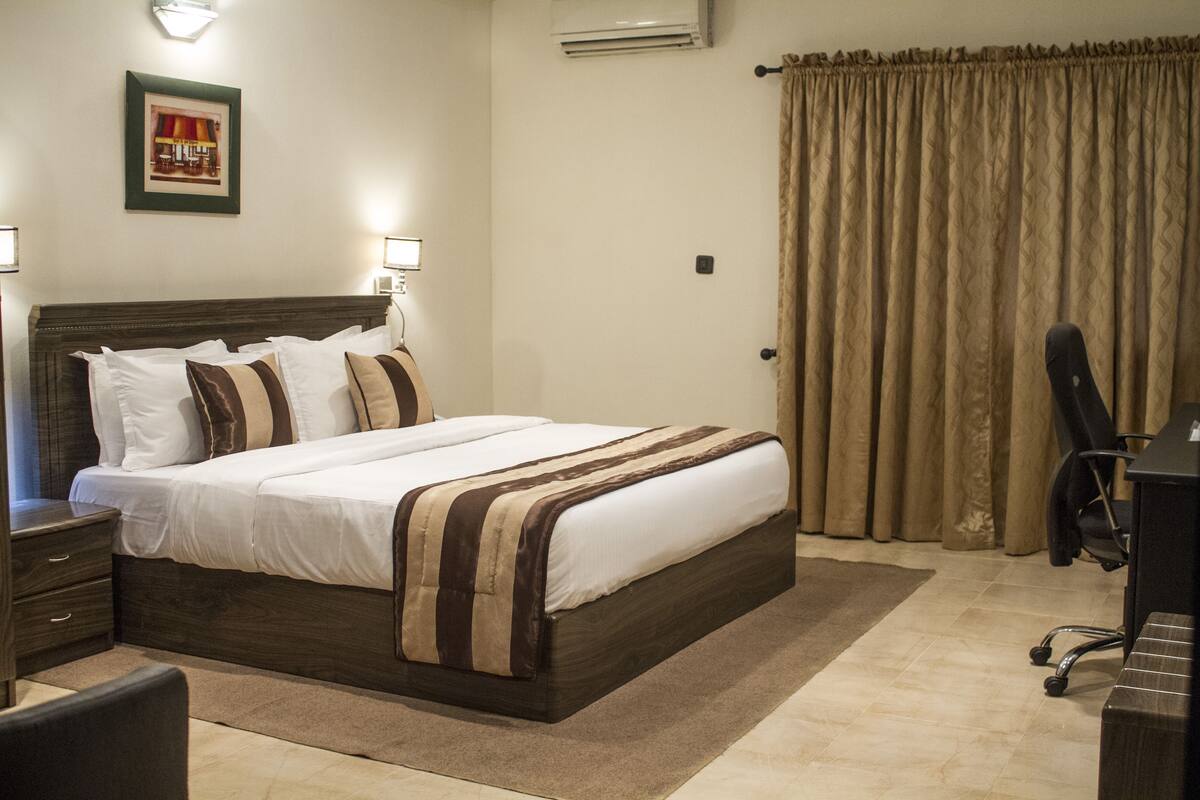 A neatly made bed with a wooden headboard, a bedside table with a lamp, and a chair in a room with beige walls and curtains.
