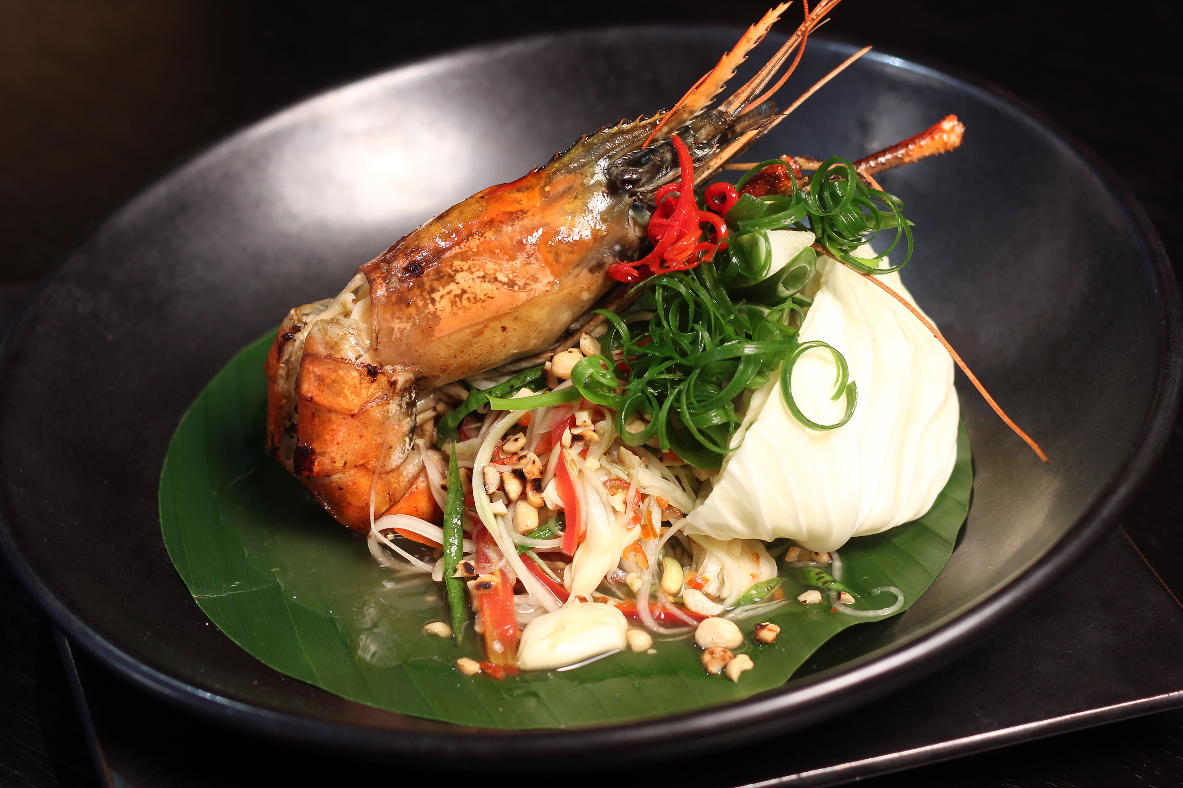A plate with a cooked shrimp, vegetables, and a white item, possibly a dumpling, on a dark plate.