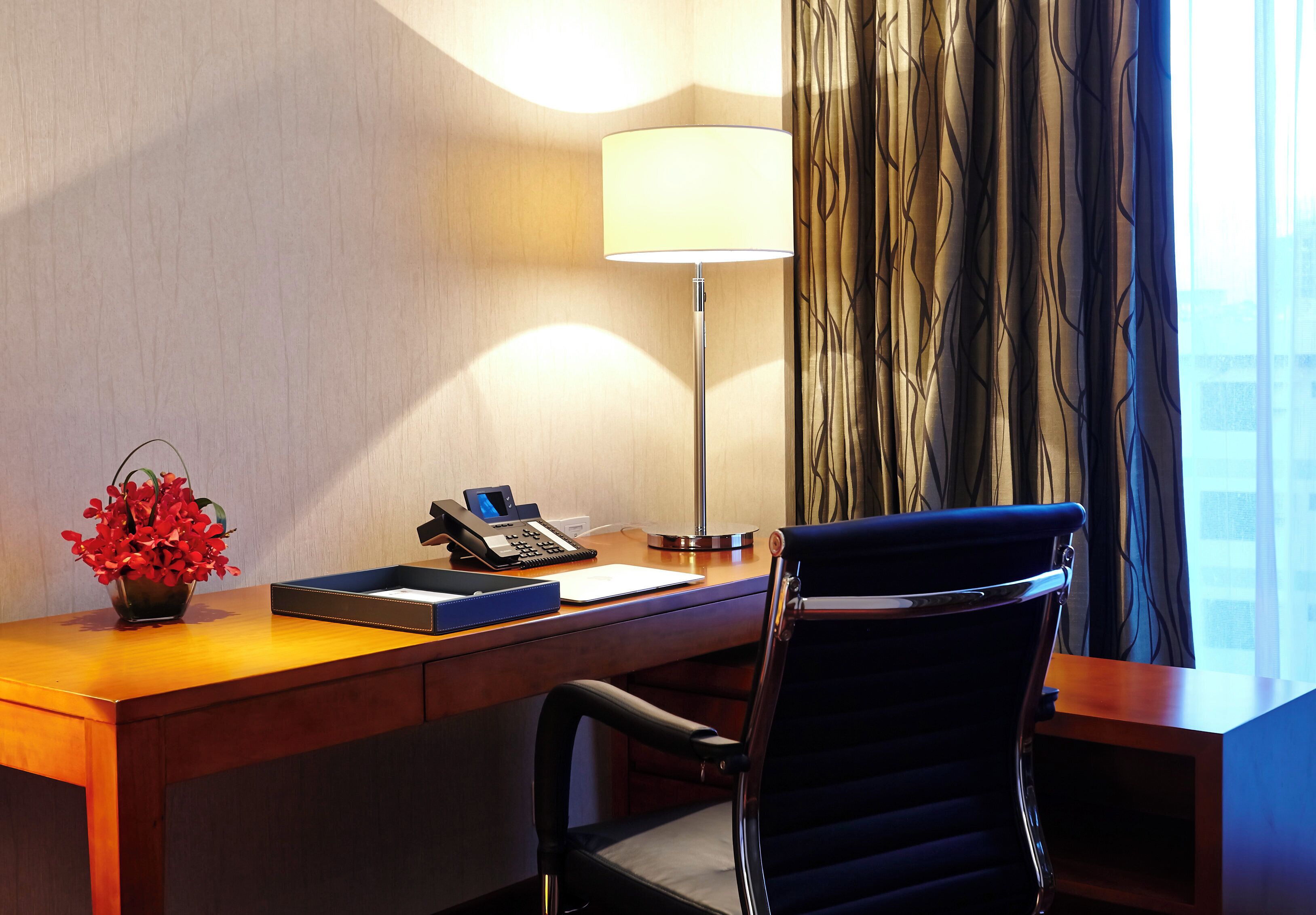 Habitación de hotel con un escritorio de madera, una lámpara, un teléfono, una silla y un florero con flores rojas.