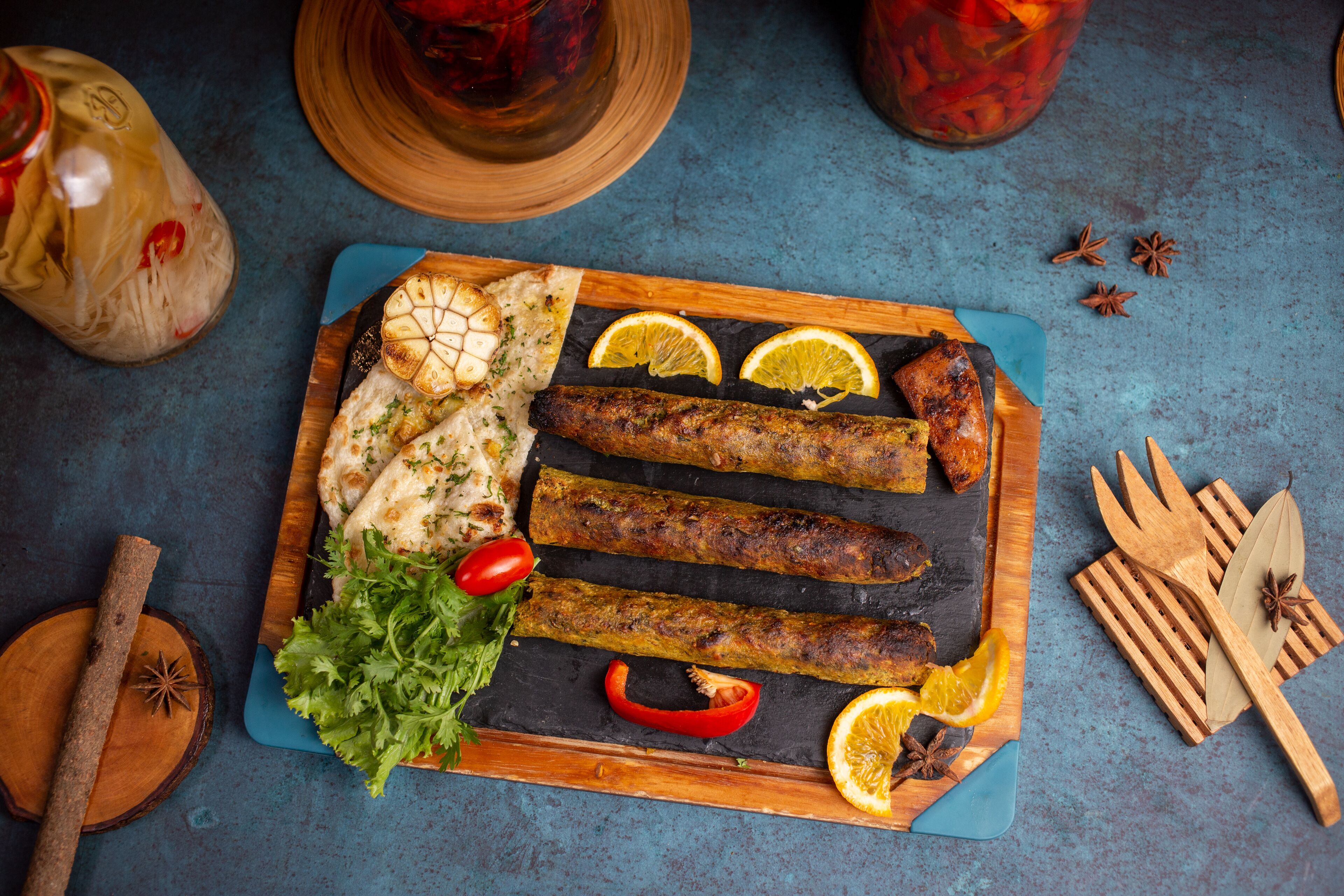 A wooden serving board with grilled meat, flatbread, and garnishes on a blue surface.