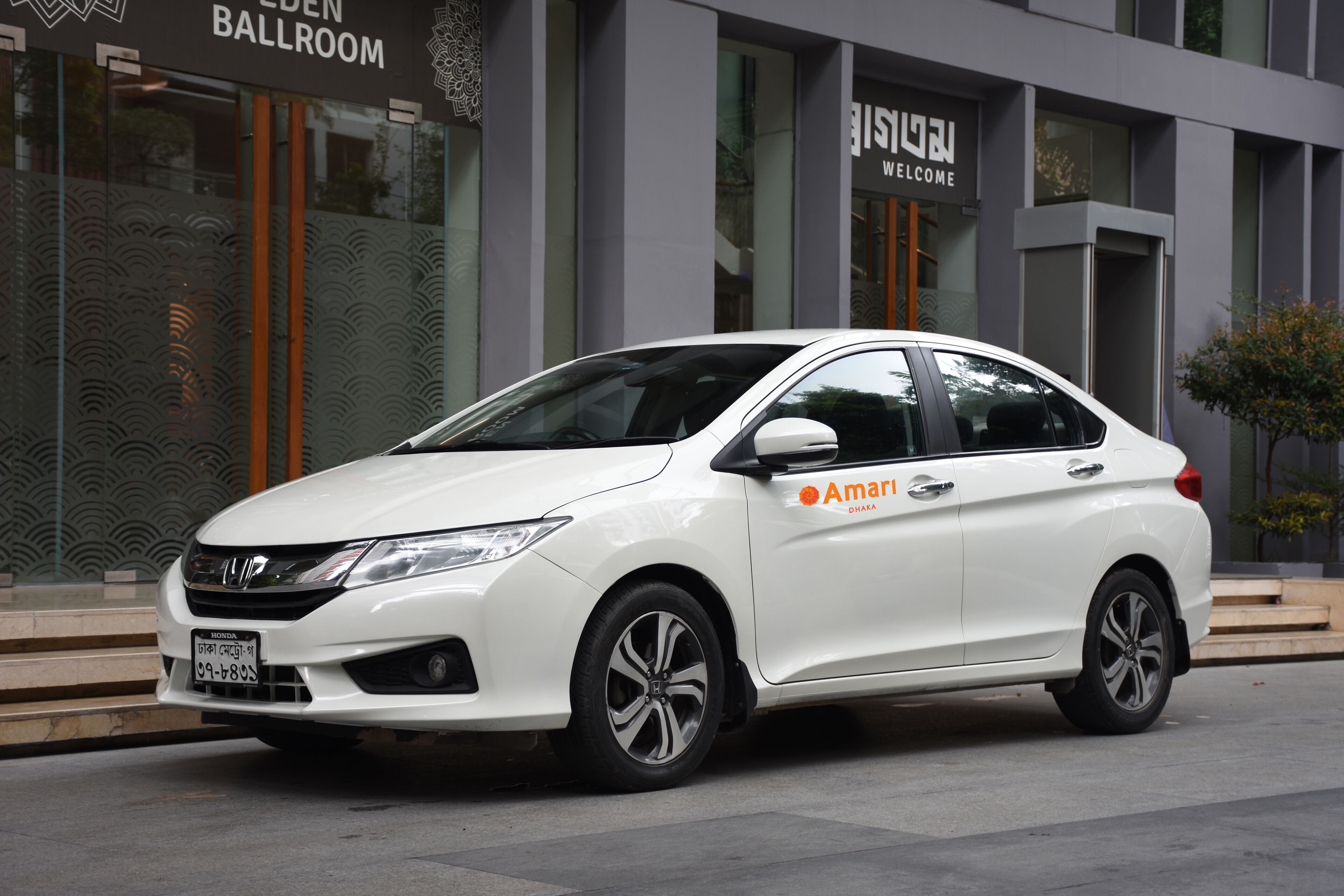 Ein weißer Honda parkt vor einem Gebäude. Ein Schild zeigt das Wort "Ballroom", ein weiteres Schild trägt die Aufschrift "Welcome" in einer anderen Sprache.