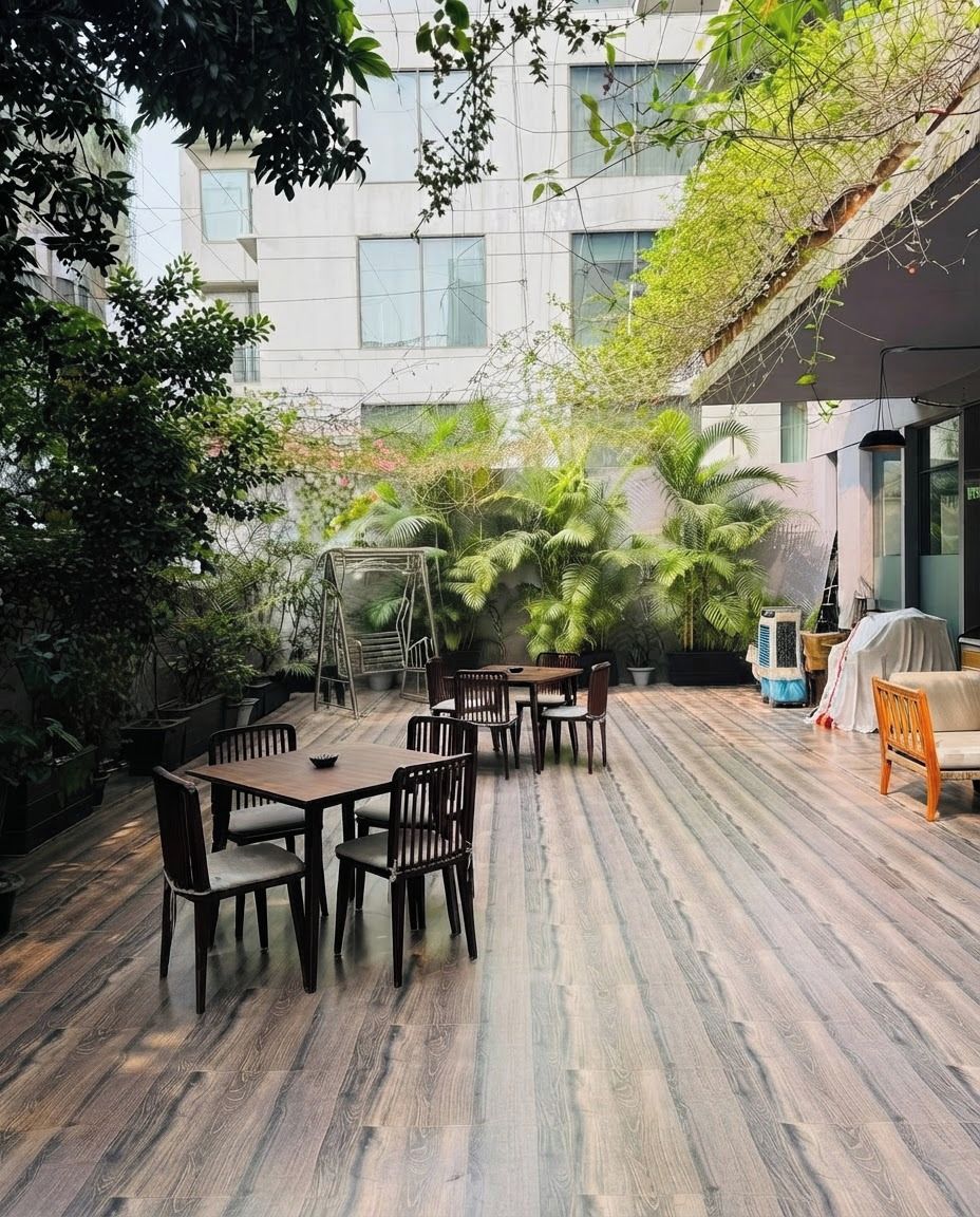 Un área de comedor al aire libre con piso de madera, mesas y sillas, rodeada de exuberante follaje y un edificio al fondo.