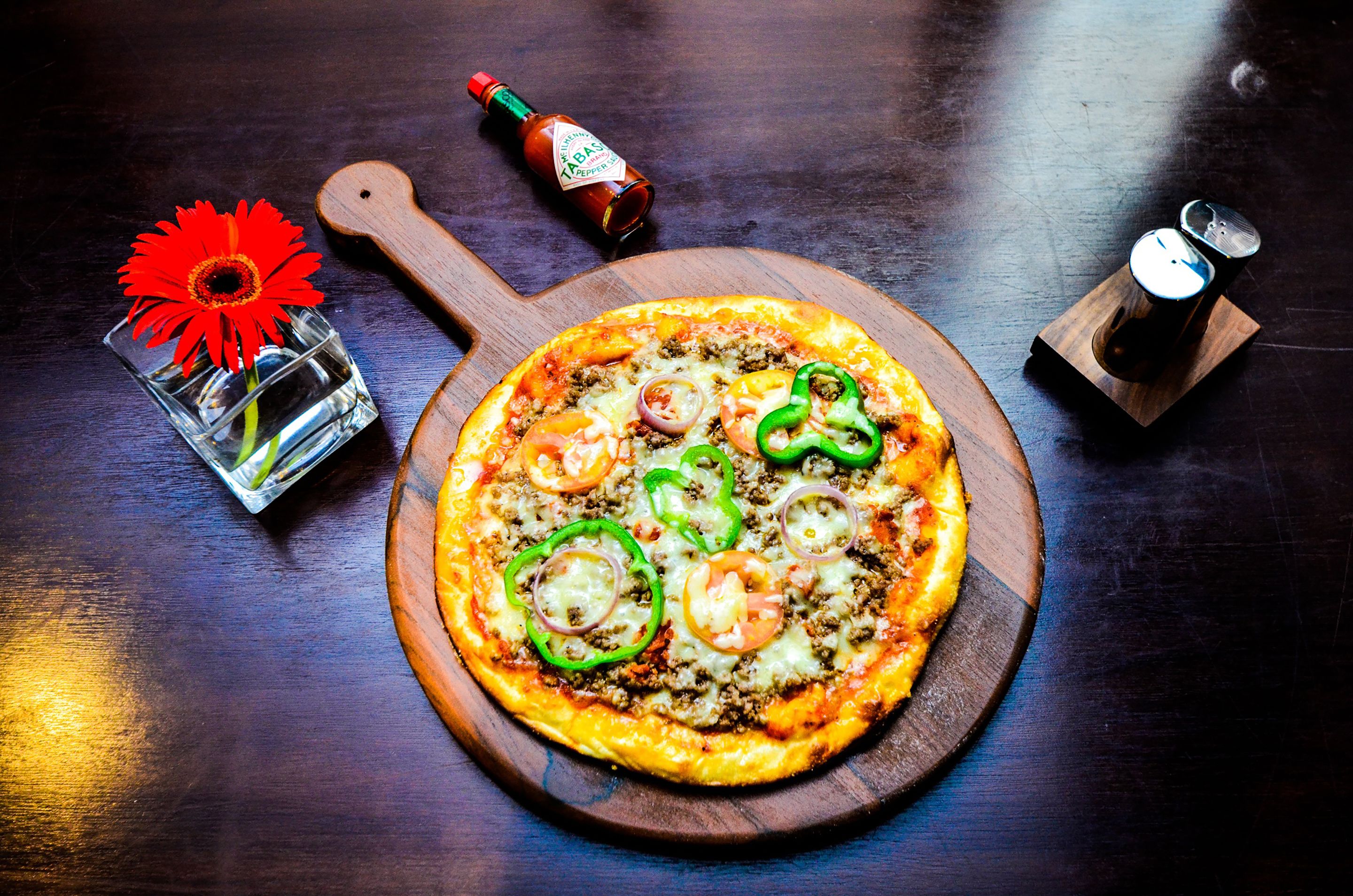Une pizza servie sur une planche en bois, accompagnée d’une bouteille de sauce Tabasco et d’un petit verre d’eau, avec une fleur à côté.