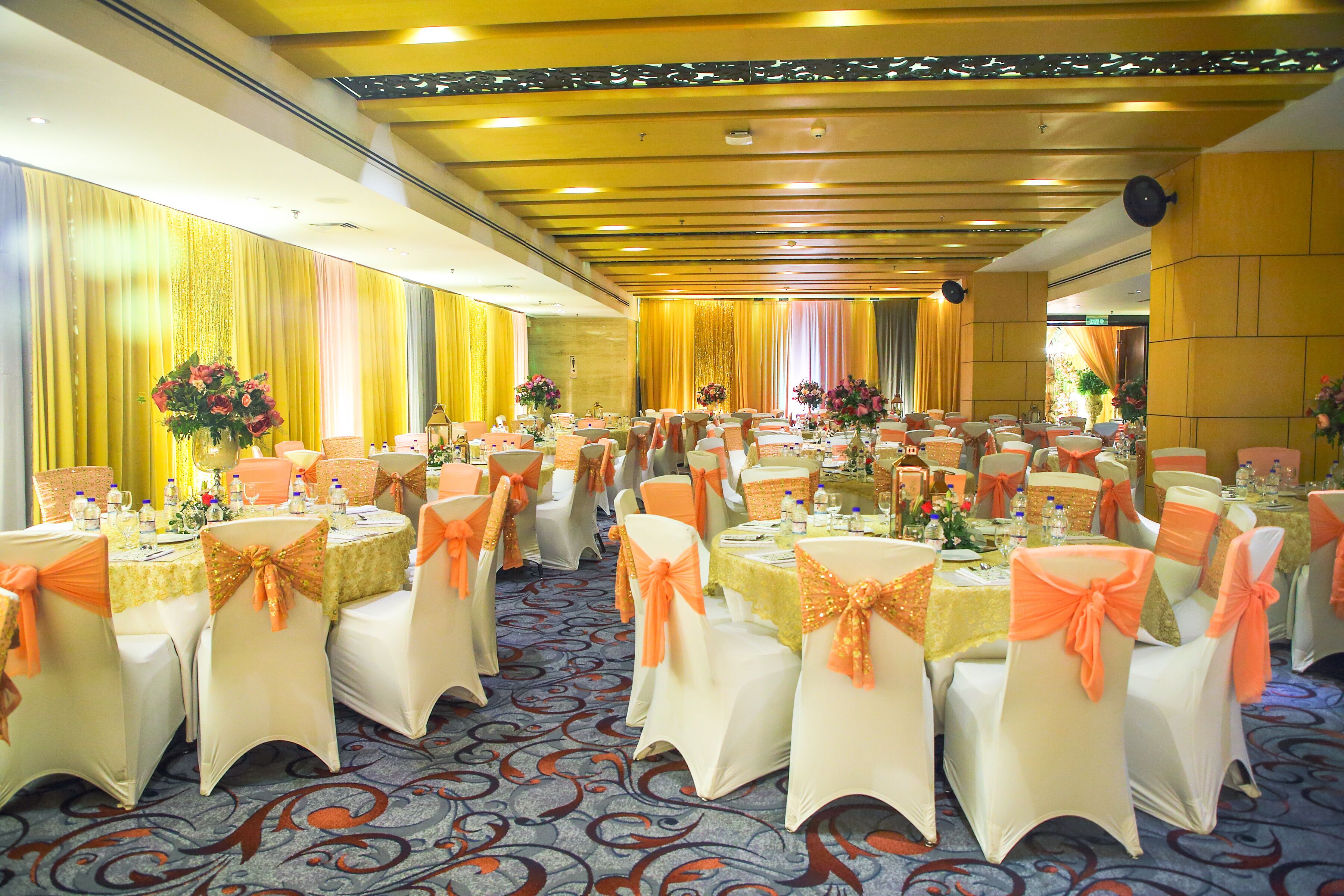 A banquet hall with round tables, white chairs with orange sashes, and floral centerpieces.