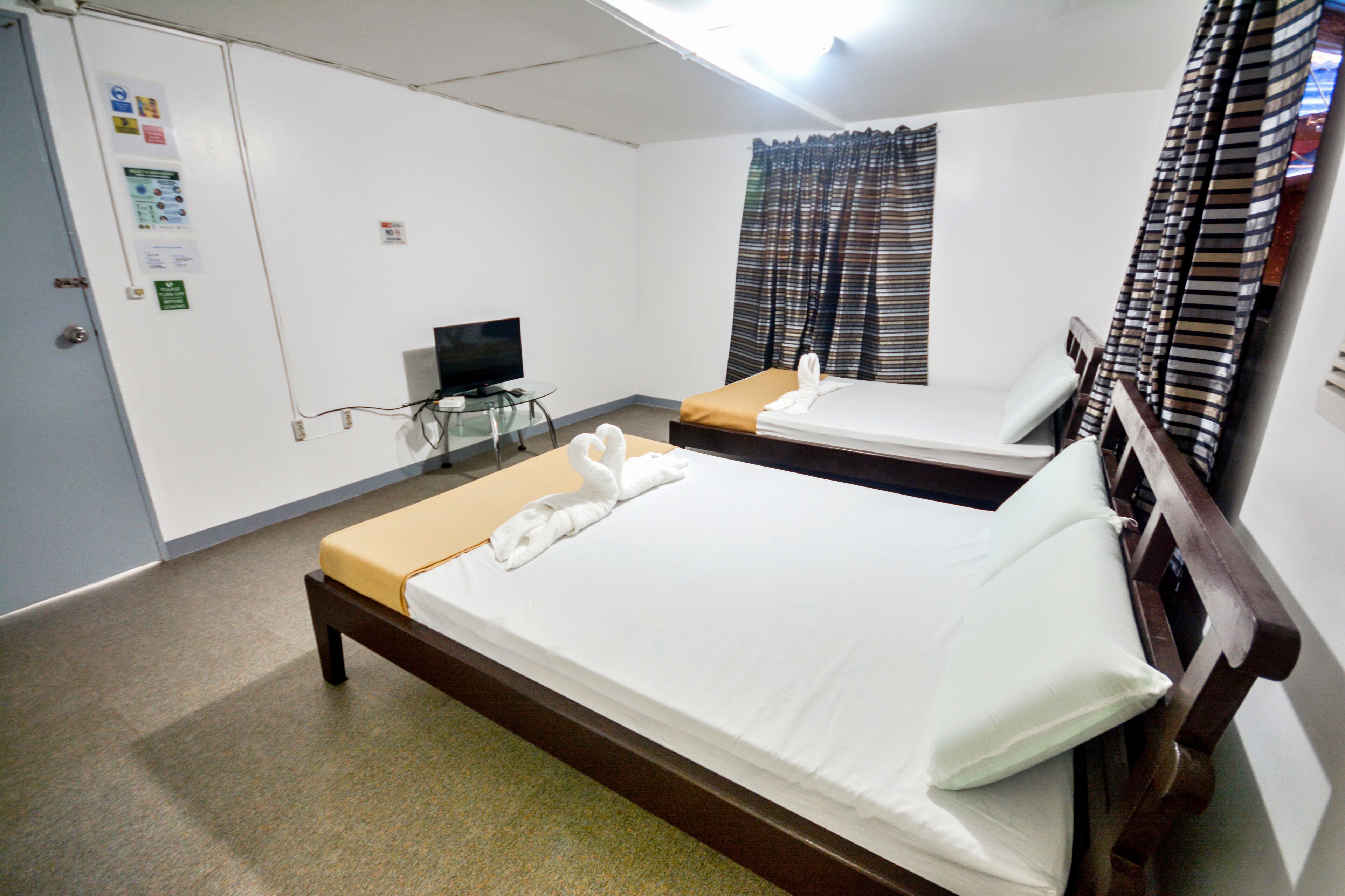 A hotel room with two single beds, a television, a small table, and a window with curtains.