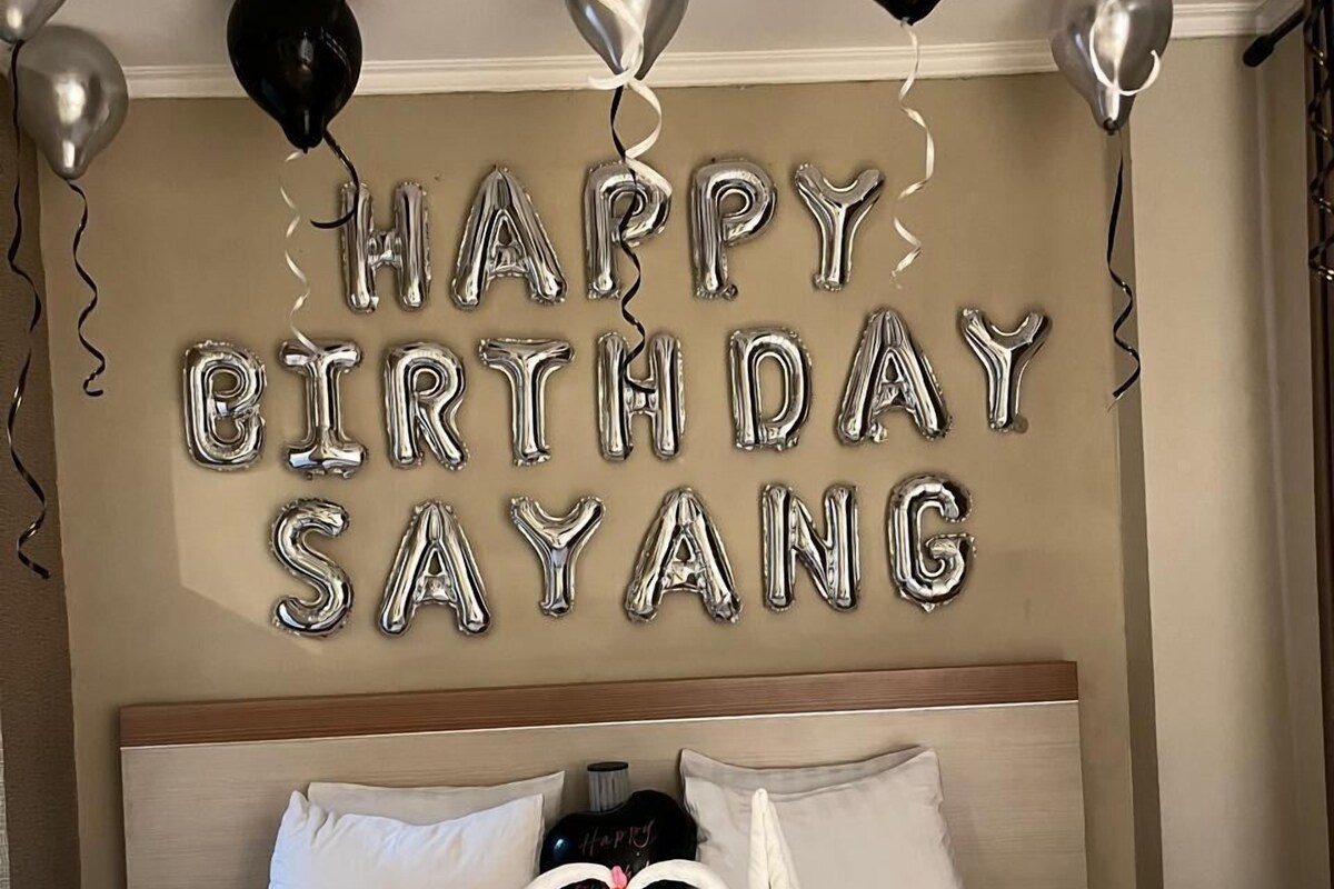 Ein Hotelzimmer mit einem Bett, Kissen, einer dekorativen Handtuchschwan und Ballons, die den Schriftzug "HAPPY BIRTHDAY SAYANG" an der Wand zeigen.