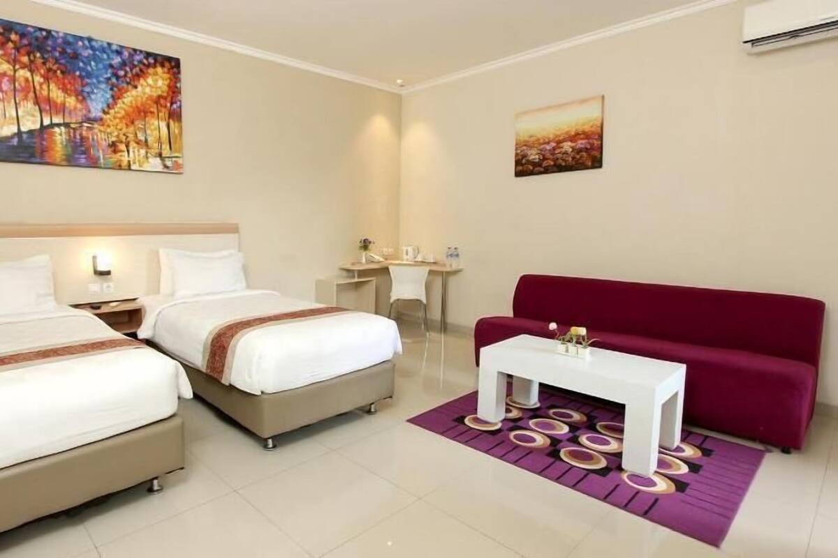 Une chambre d’hôtel avec deux lits, un canapé rouge, une petite table blanche et deux tableaux accrochés aux murs.