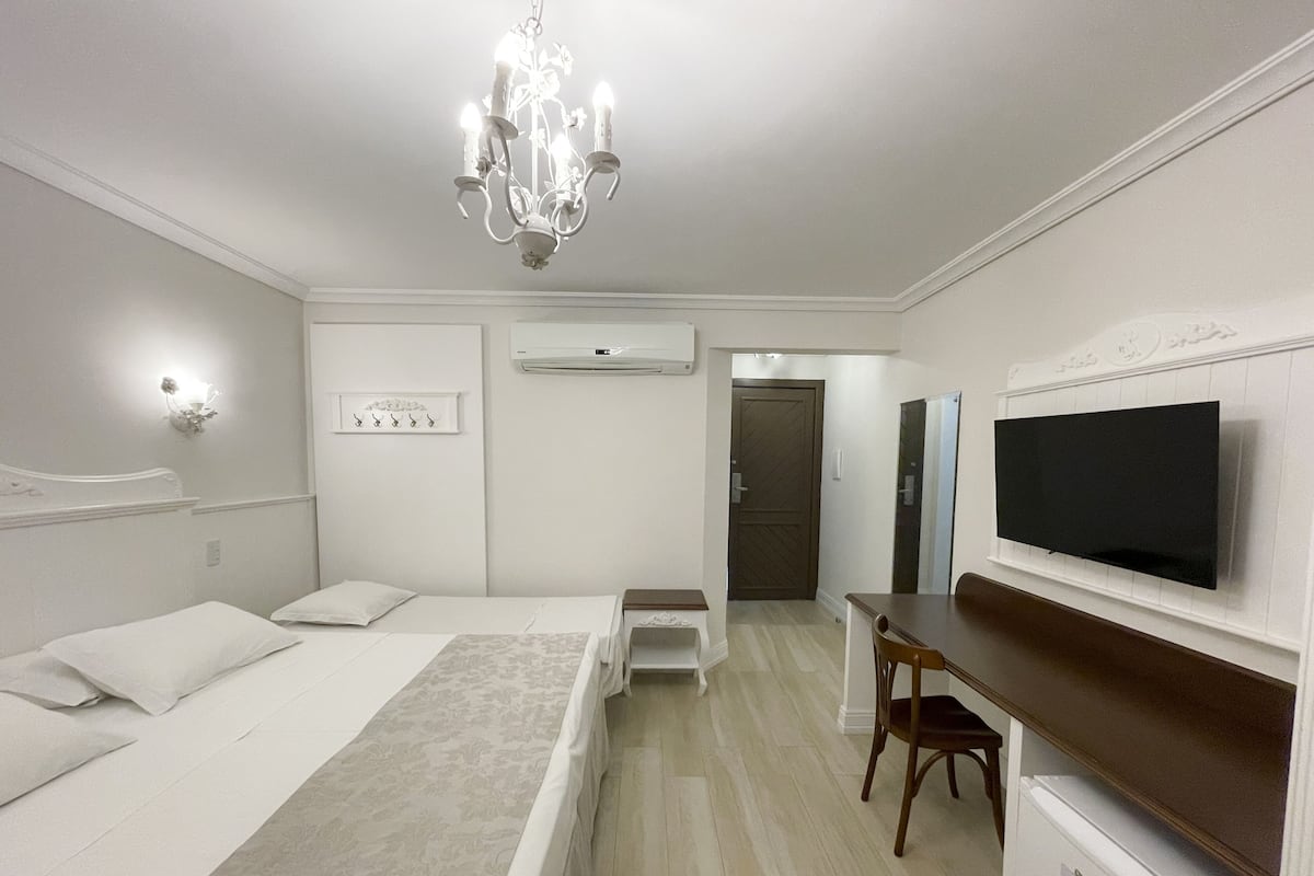A hotel room with a bed, bedside table, desk, chair, TV, and a chandelier.
