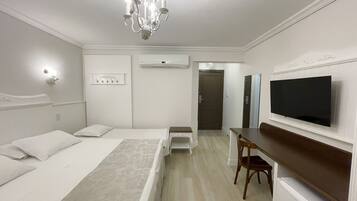 Quarto de hotel com cama, mesa de cabeceira, escrivaninha, cadeira, televisão e um candelabro.