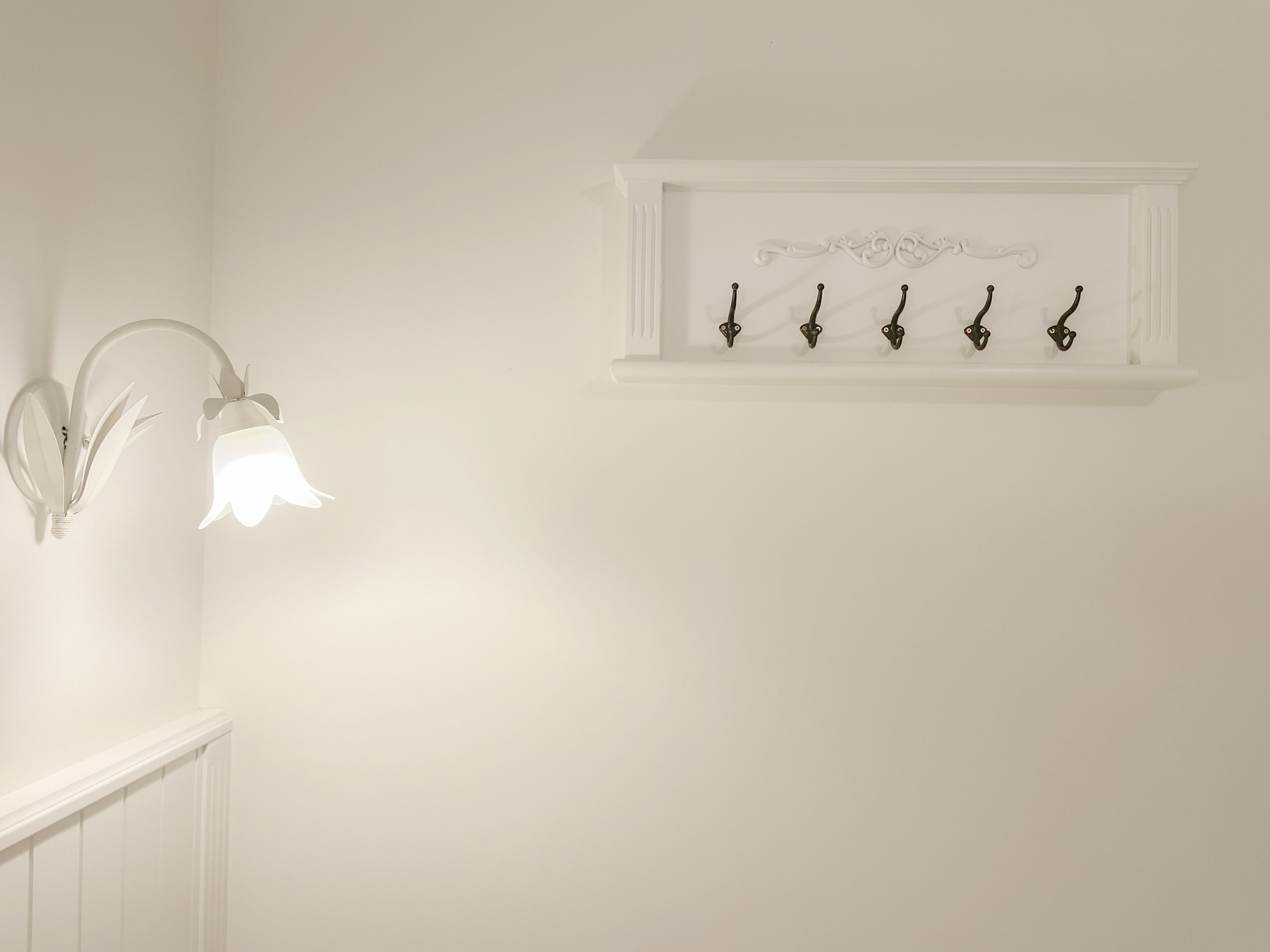 En vit vägg med en väggmonterad lampa och en hylla med fem krokar.