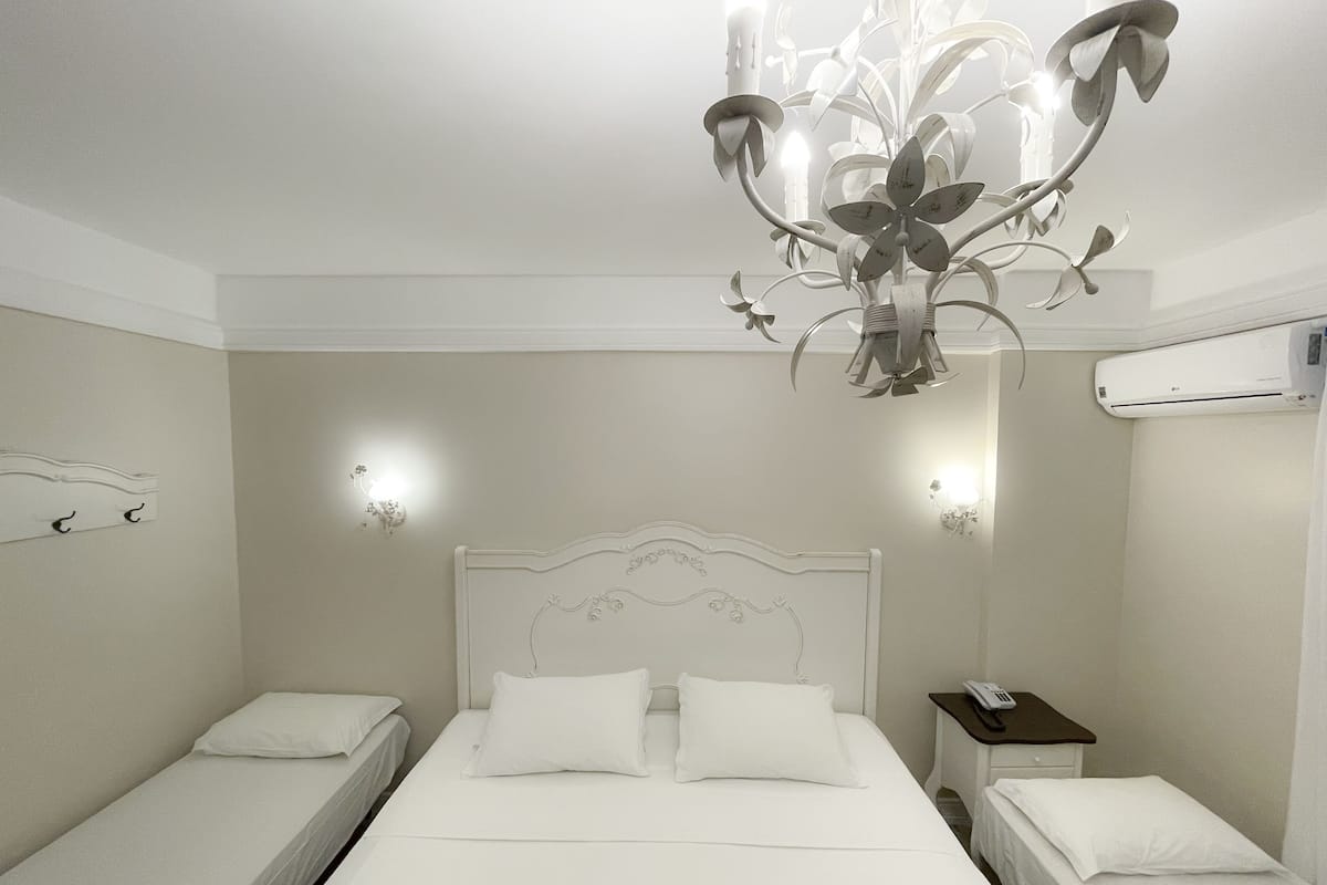 A hotel room with a large bed, two smaller beds, a chandelier, wall-mounted lights, an air conditioning unit, and a bedside table with a phone.
