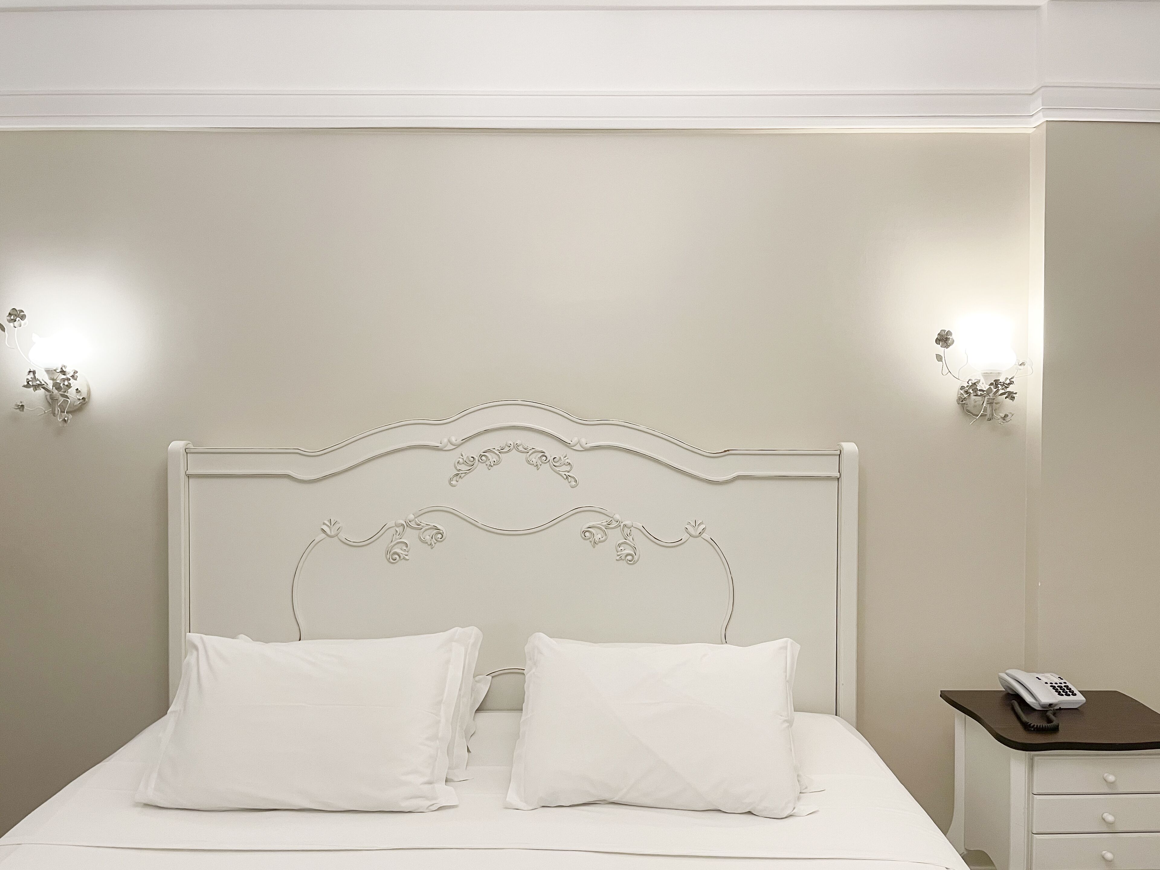 Uma cama bem arrumada com roupa de cama e almofadas brancas, uma cabeceira branca com detalhes decorativos em relevo e uma mesa de cabeceira com um abajur e um telefone.