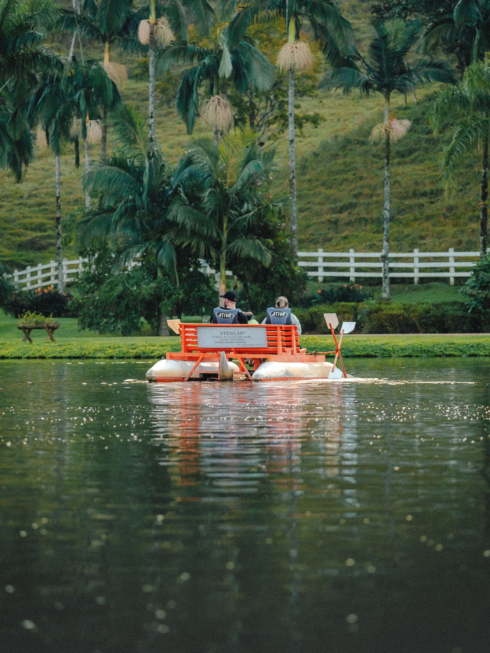 A boat with two people wearing 'ATENÇÃO' vests on a calm water body with a white fence and palm trees in the background.
