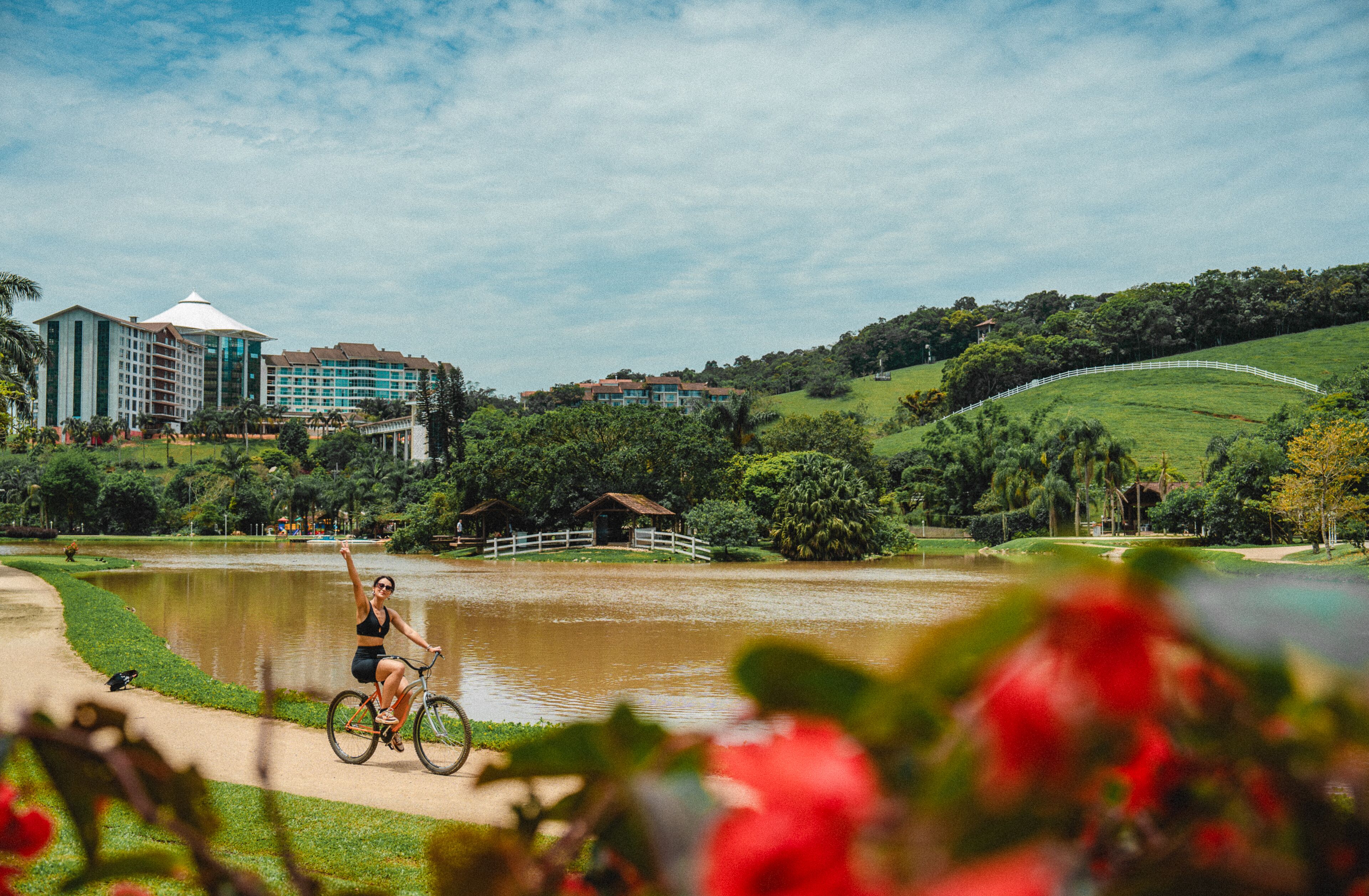 Uma pessoa andando de bicicleta em um caminho perto de um corpo d'água, com edifícios e vegetação ao fundo.