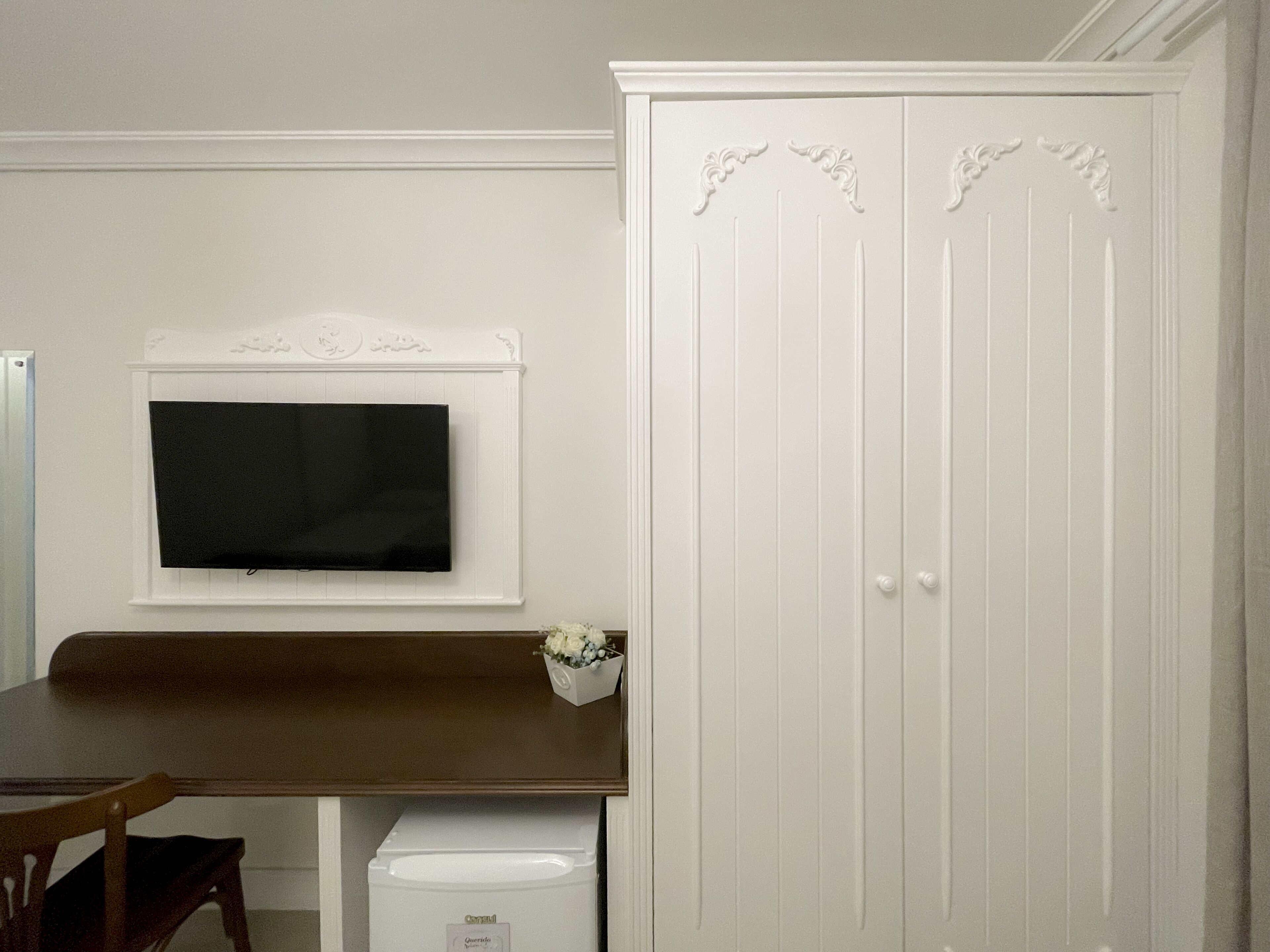 Quarto com guarda-roupa branco, televisão montada e uma pequena mesa com um vaso de flores.