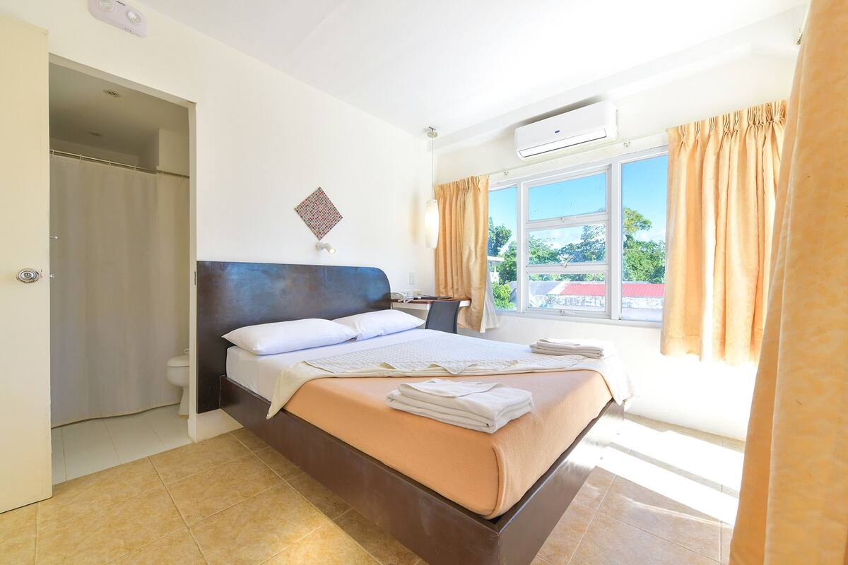 Une chambre d’hôtel équipée d’un lit, d’un bureau, d’une chaise, d’une fenêtre avec des rideaux et d’un appareil de climatisation.