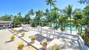 Una terraza con mesa y sillas, plantas en macetas y vistas a palmeras y una playa.