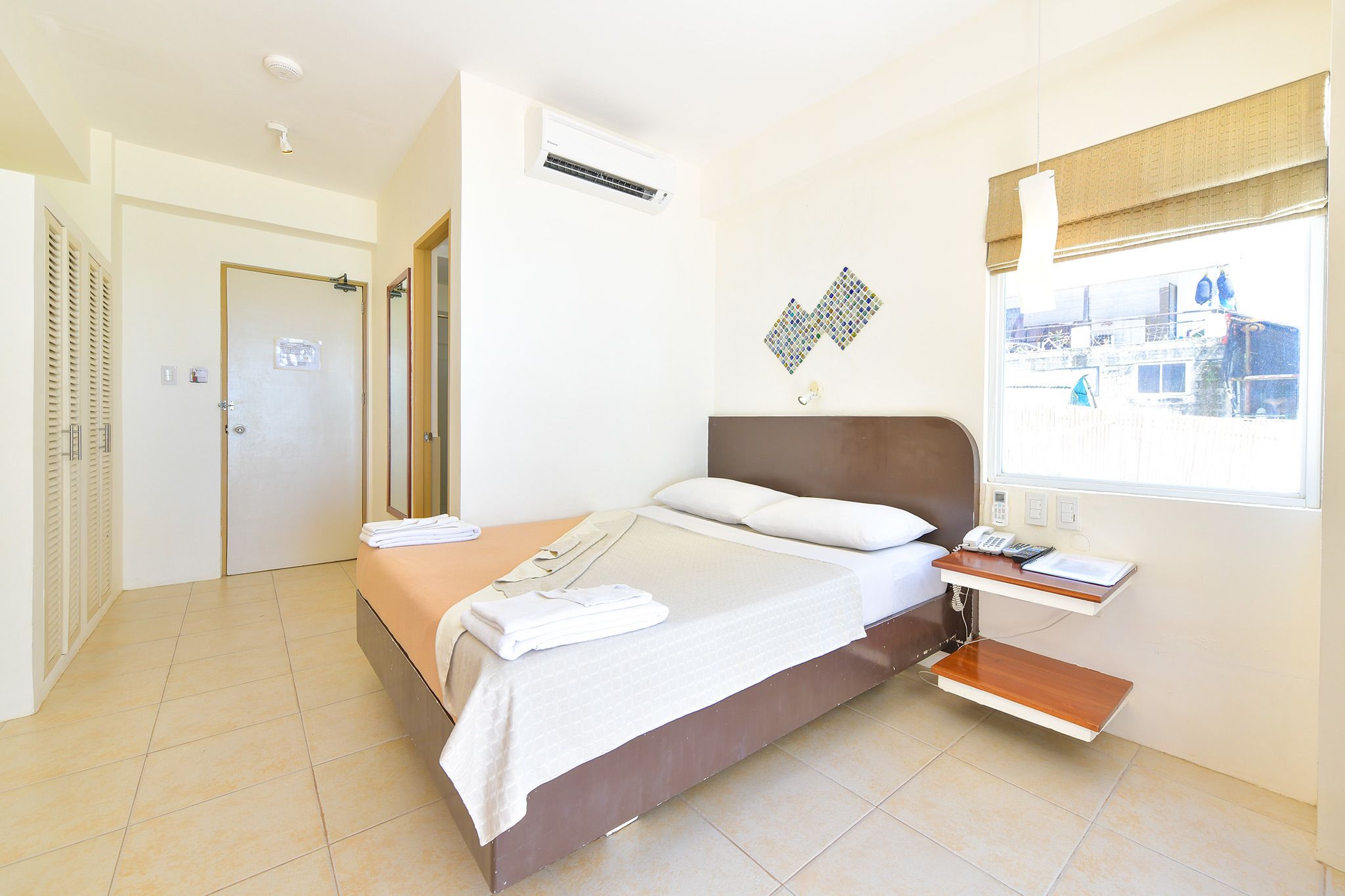 A hotel room with a bed, a bedside table, a window with a view, and an air conditioning unit.