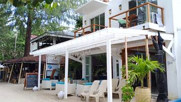 A beachside cafe with outdoor seating, a covered patio, and a two-story building with a balcony.
