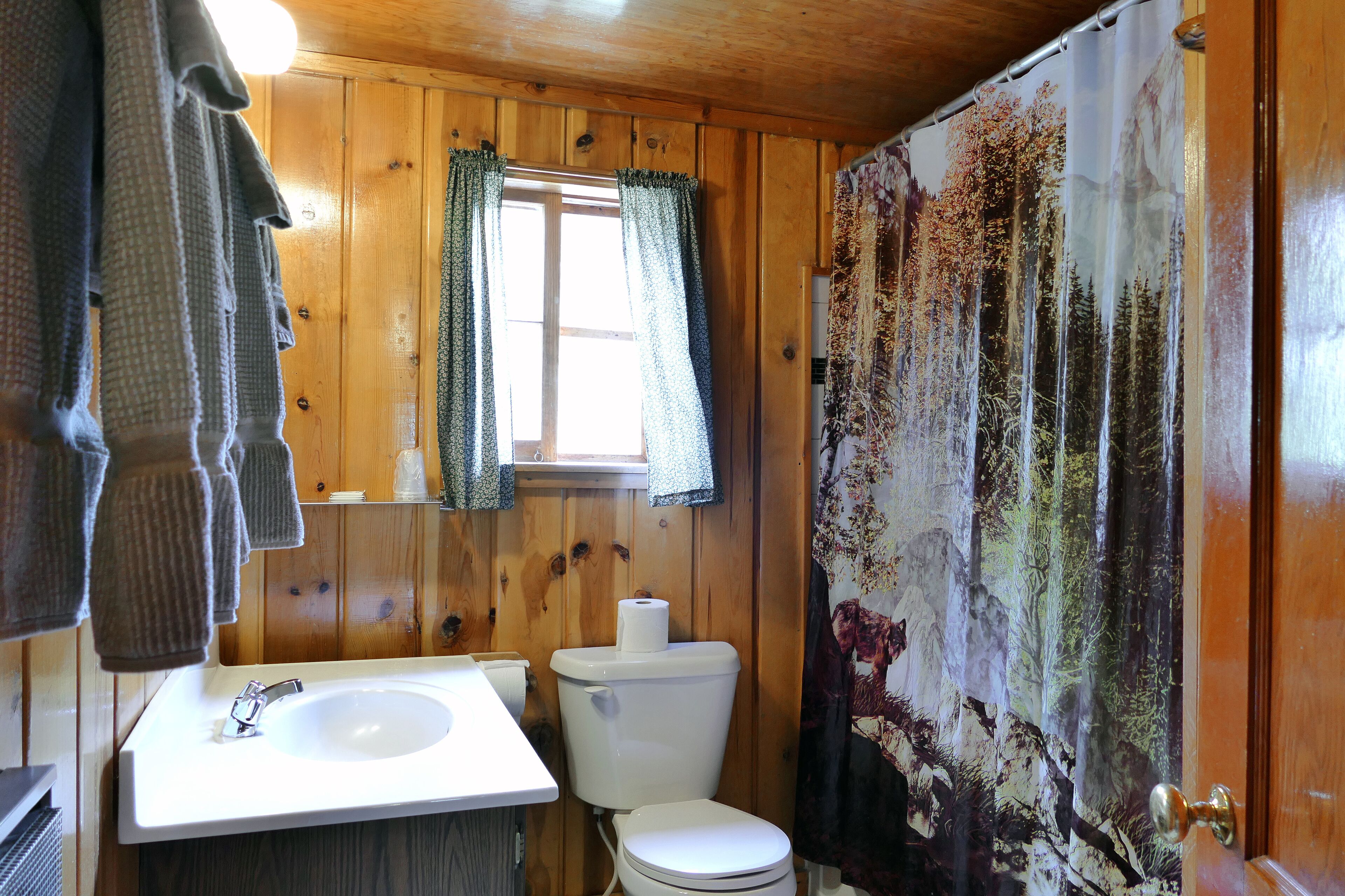 A wooden bathroom with a white sink, toilet, and shower curtain with a forest print.