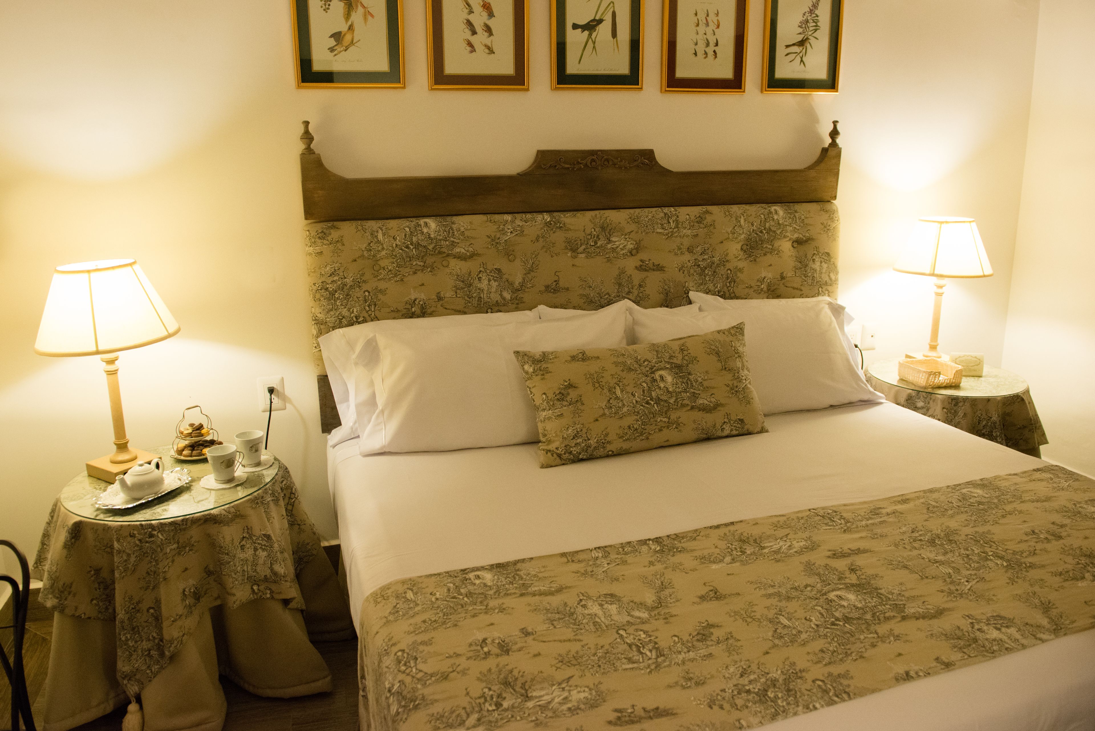 A neatly made bed with a floral bedspread and matching pillows, two bedside tables with lamps, and a wall decorated with framed botanical prints.