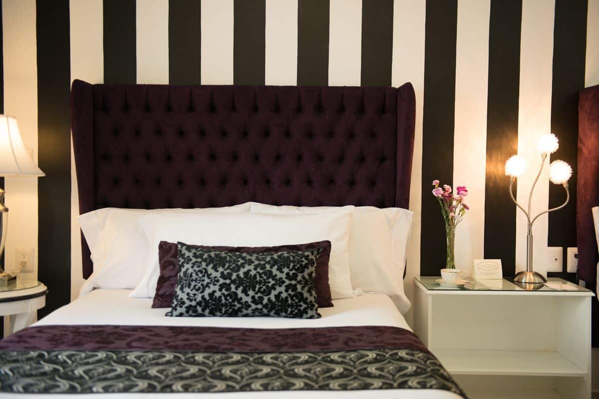 A neatly made bed with a dark headboard, white pillows, and a patterned throw blanket. There is a bedside table with a lamp and a vase with flowers.