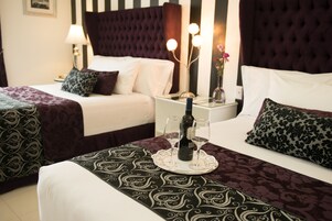 Habitación de hotel con dos camas, una mesita de noche con una botella y vasos, y un jarrón decorativo con flores.