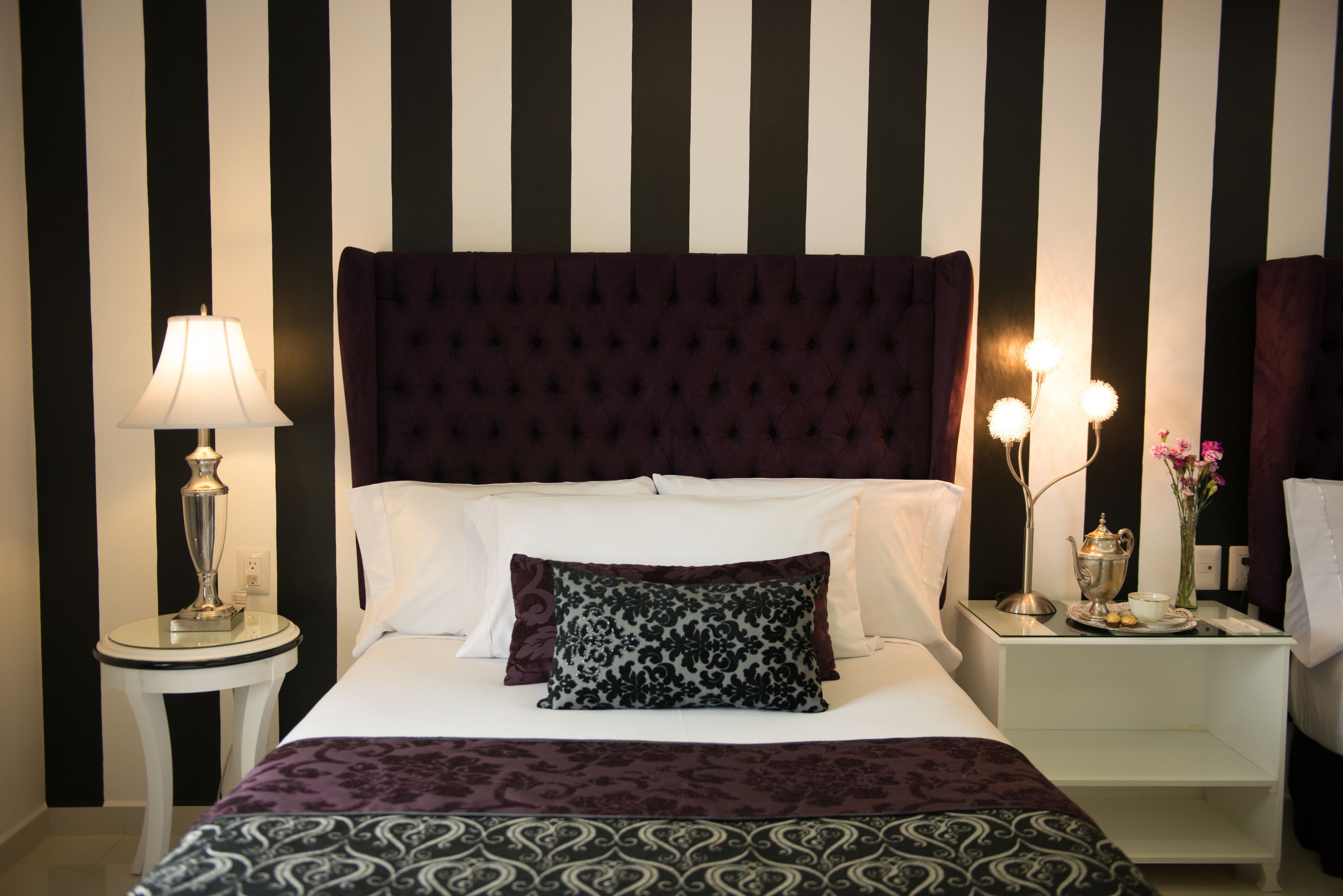 Una habitación de hotel con una pared a rayas, un cabecero capitonado, una mesita de noche con una lámpara y un pequeño mueble con un juego de té.
