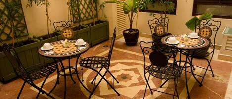 Un patio con una mesa redonda y sillas, rodeado de macetas con plantas y un muro decorativo.