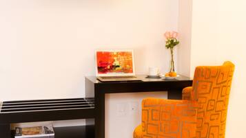 A modern hotel room with a flat-screen TV mounted on the wall, a laptop on a desk, a magazine, a vase with flowers, and an orange patterned chair.