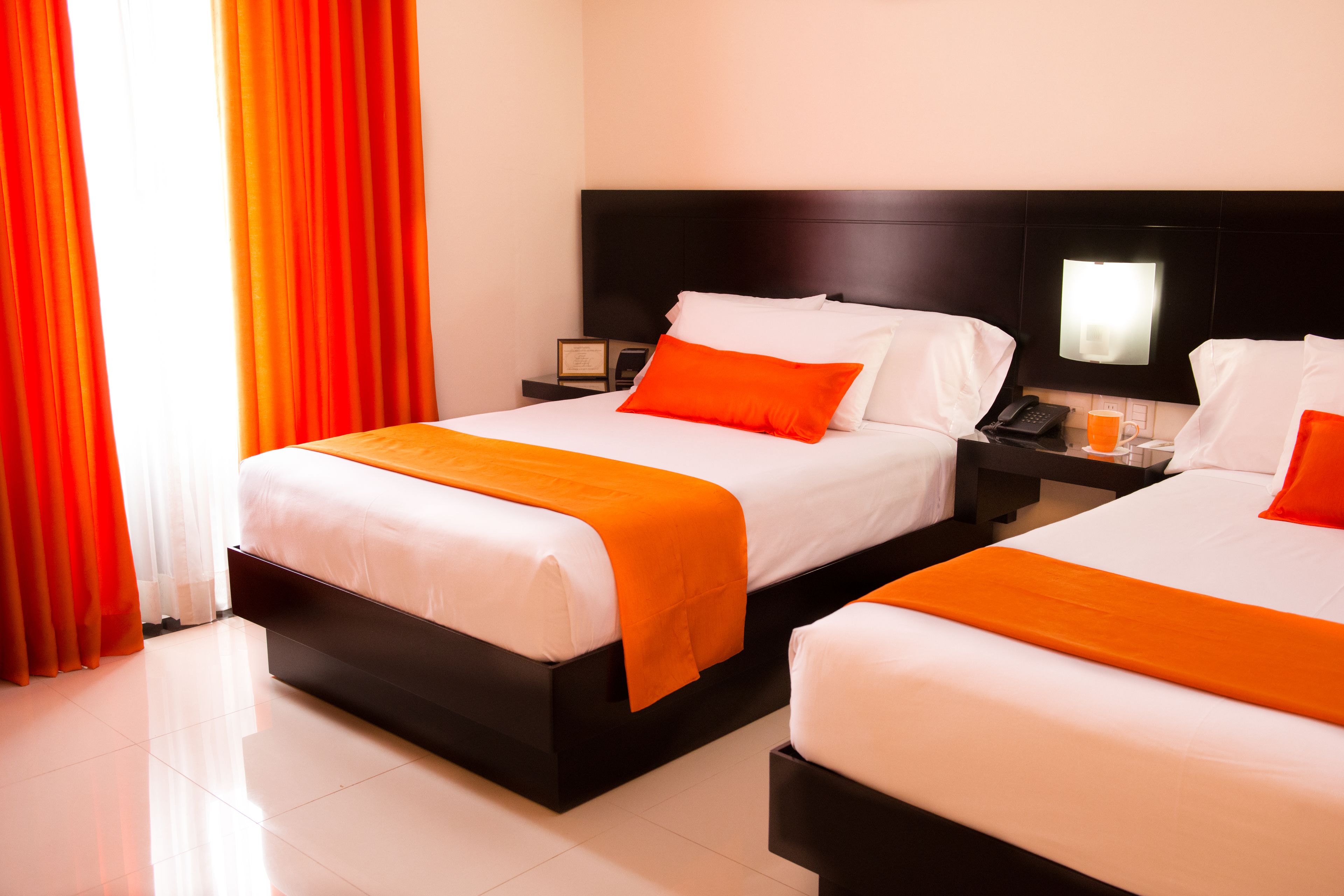 Habitación de hotel con dos camas, cada una con ropa blanca y detalles naranja, una mesita de noche con un teléfono y una taza, y un ventanal con cortinas.