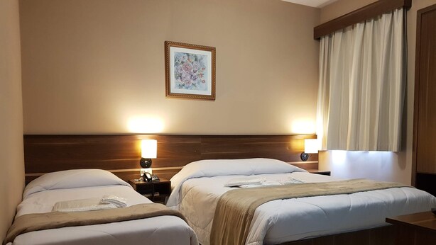 A hotel room with two beds, a nightstand with a lamp, a framed picture on the wall, and a window with curtains. - Astro Hotel (São José dos Campos)