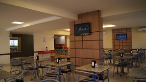 A spacious restaurant with multiple tables and chairs, a television mounted on the wall, and a counter area with a red tablecloth. - Astro Hotel (São José dos Campos)
