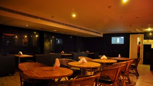 A dimly lit indoor space with round tables and wicker chairs, a television screen, and a door with a sign. - Nayagara Hotel (Chennai)