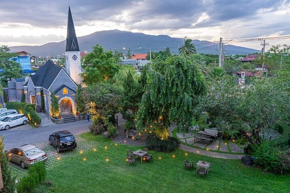 A church with a steeple, surrounded by lush greenery and a parking area.