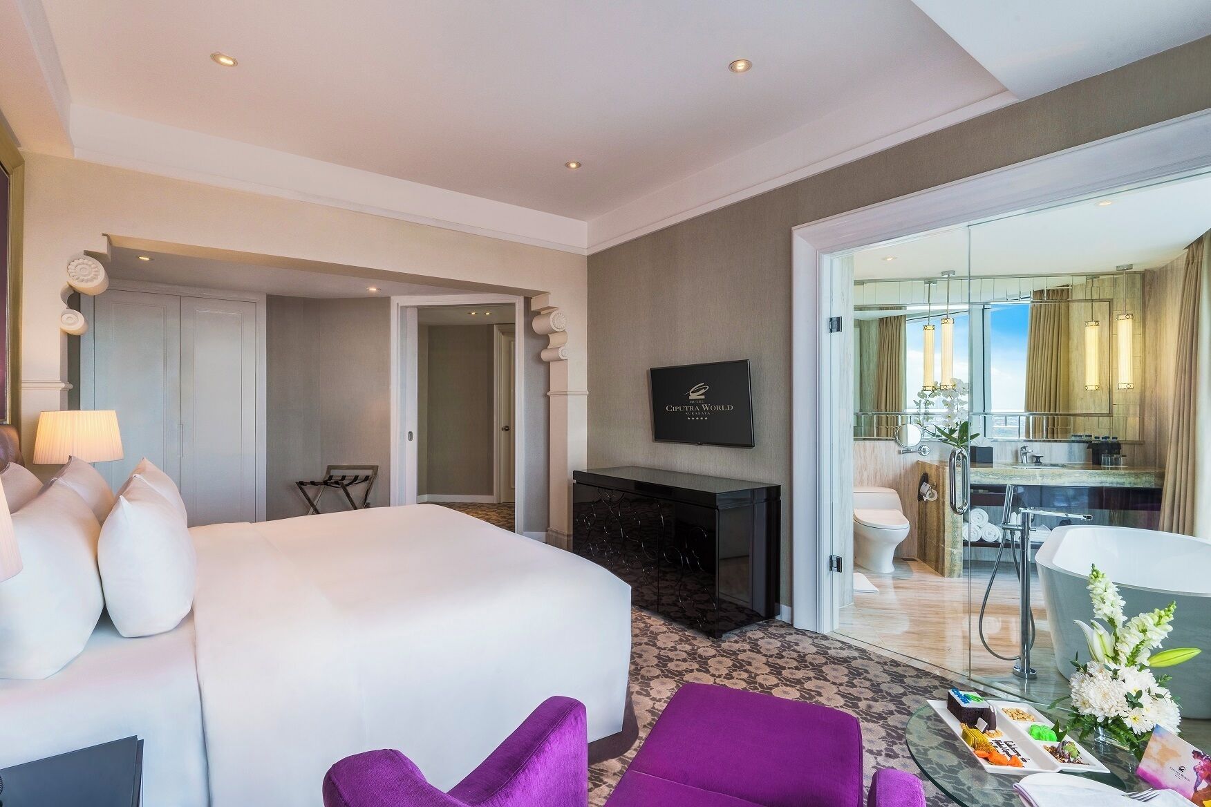 A hotel room with a large bed, a TV, a desk, and a view of the kitchen and balcony.