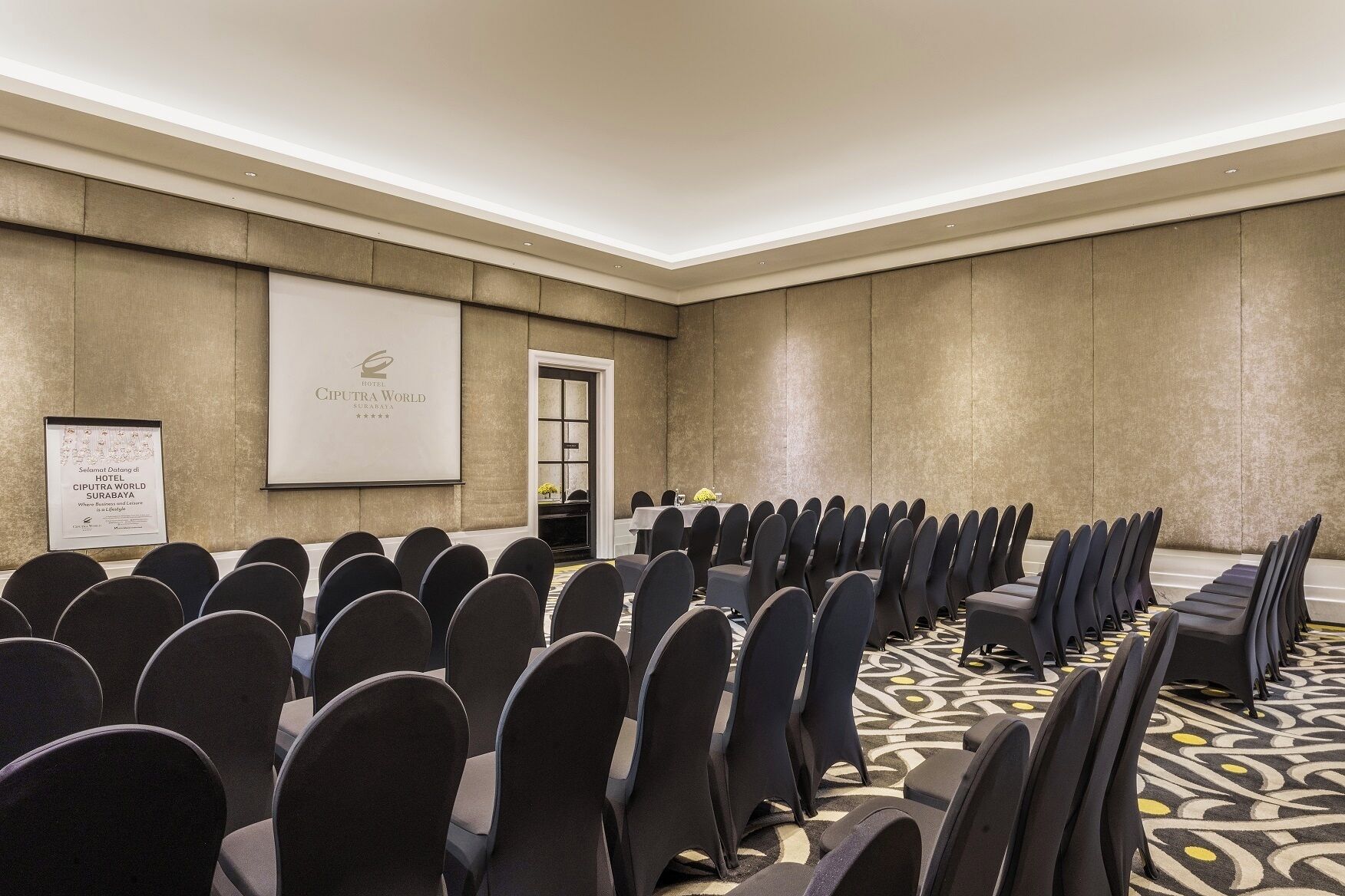 A conference room with rows of chairs, a projector screen, and a sign for Hotel Ciputra World Surabaya.