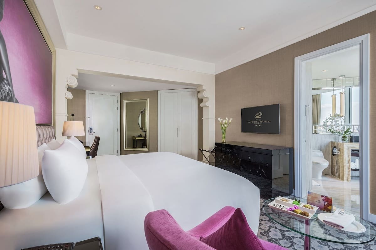 A modern hotel room with a large bed, a sitting area with a chair, a TV, a fireplace, and a dining area with a table and plates.