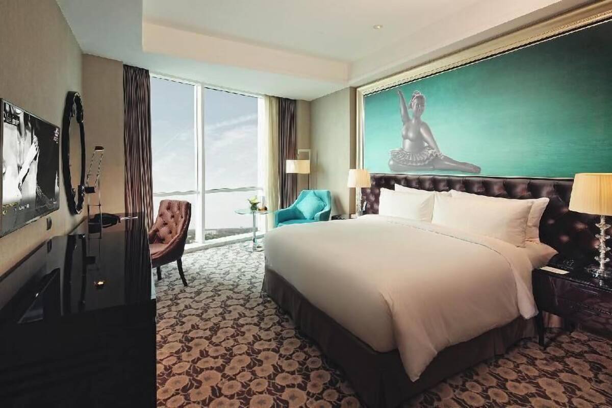 A hotel room with a large bed, a TV, a chair, a painting, and a view of the outdoors.