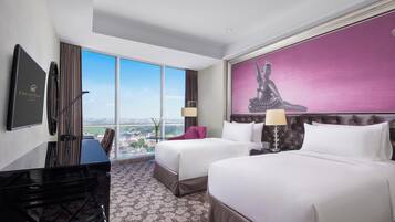 A hotel room with two beds, a desk, a large window with a city view, and a painting on the wall.