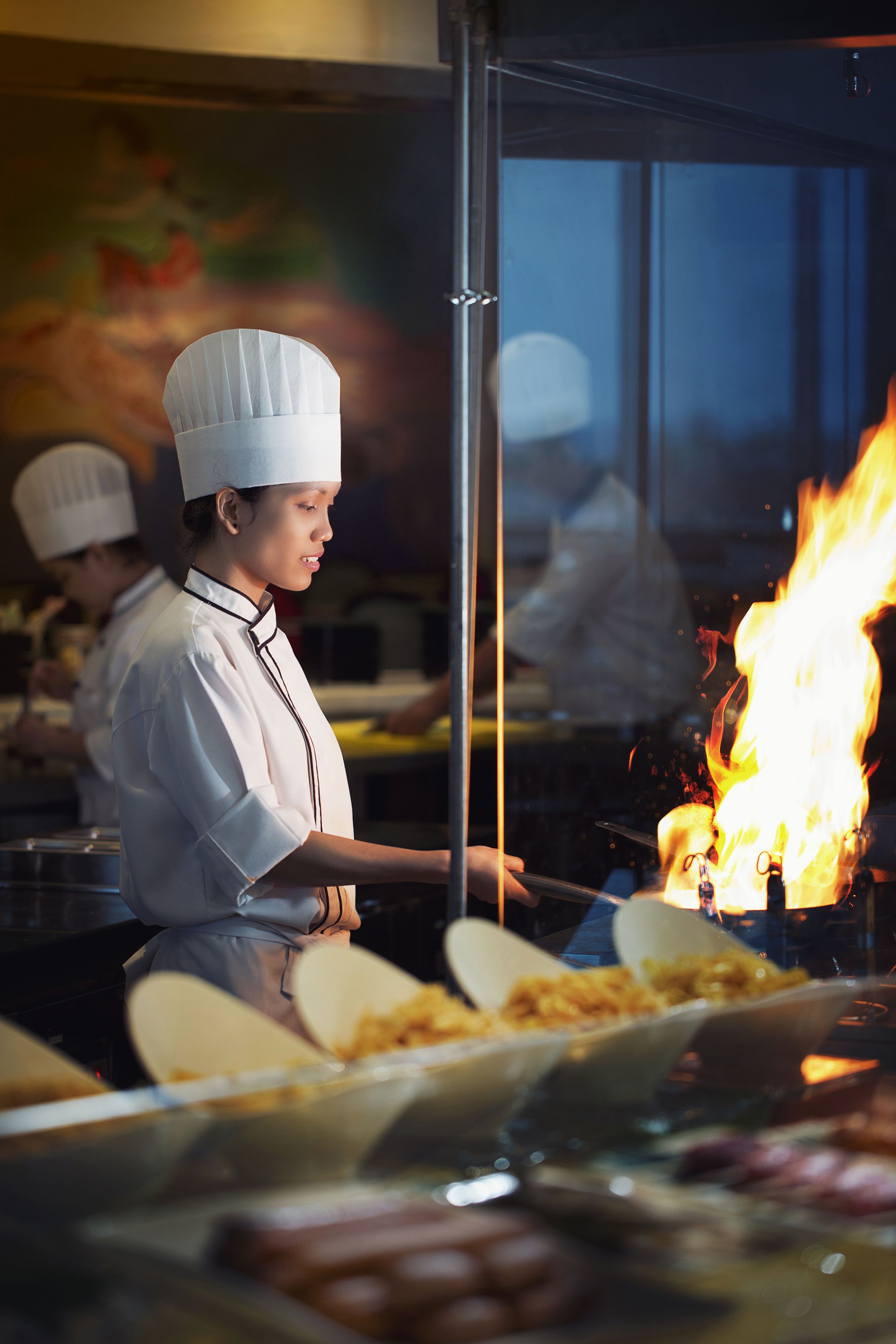 Un chef con uniforme blanco y gorro cocina, con llamas visibles al fondo.