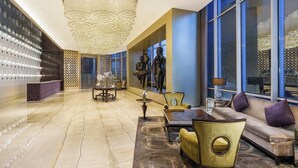 A spacious lobby with a marble floor, a large chandelier, and a statue of a dancer. There is a reception desk, a dining area, and a seating area with a sofa and chairs.