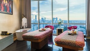 A modern hotel room with a large city view, two massage tables, a bathtub, and a cityscape visible through large windows.