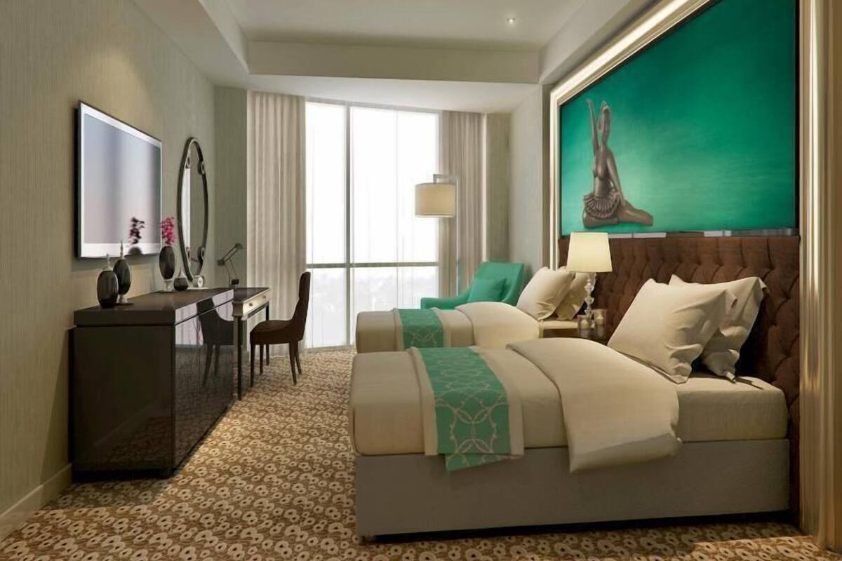 A hotel room with a large bed, a desk, a chair, a large window, and a painting on the wall.