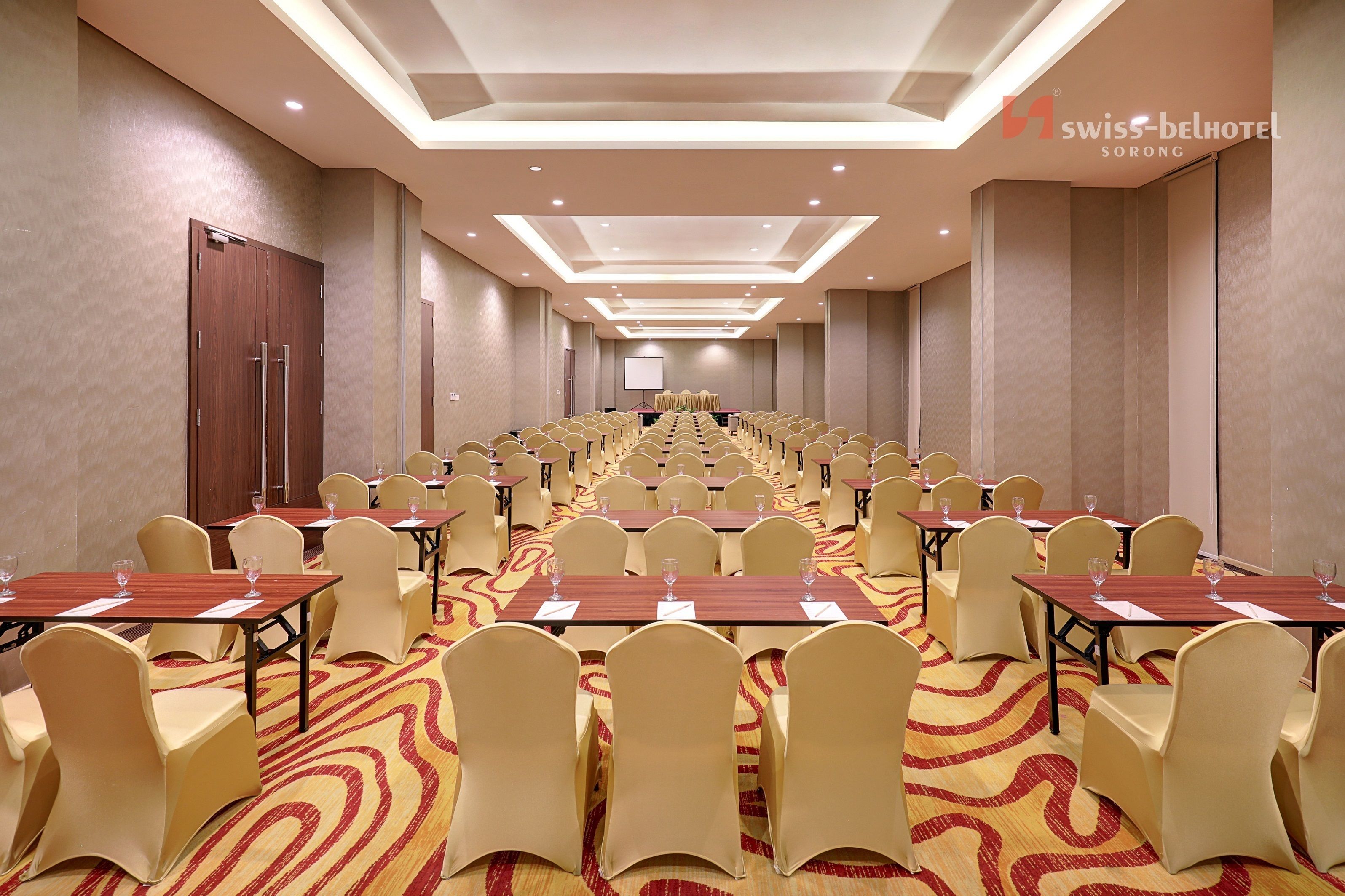 A conference room with rows of tables and chairs, a large screen at the front, and a carpeted floor.