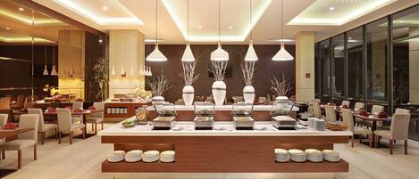 A modern hotel lobby with a central buffet, multiple dining tables, and a large window providing a view of the city.