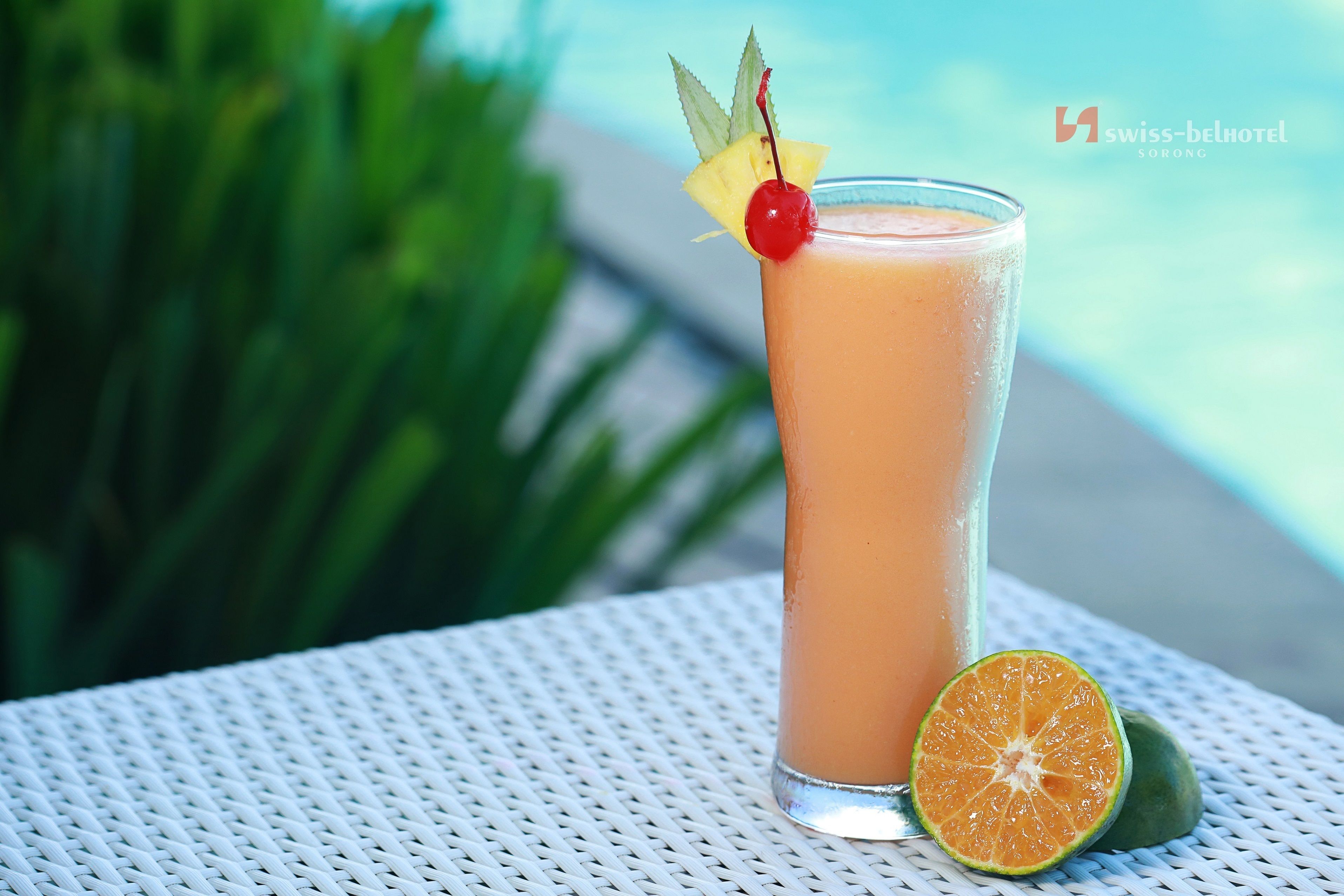 A tall glass of orange-colored drink with a cherry and pineapple garnish, placed on a white woven table with a green plant in the background.