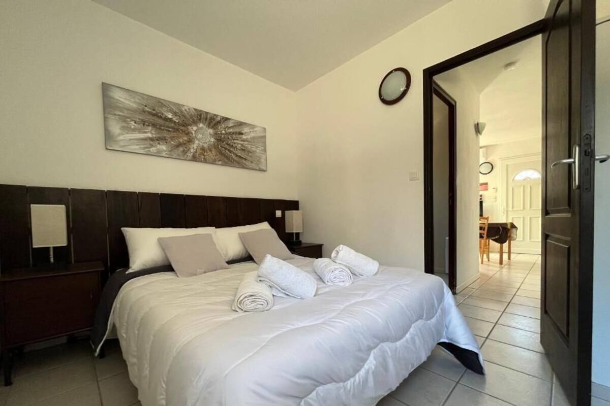 A neatly made bed with white linens and rolled towels, a wooden headboard, and a wall-mounted abstract painting.