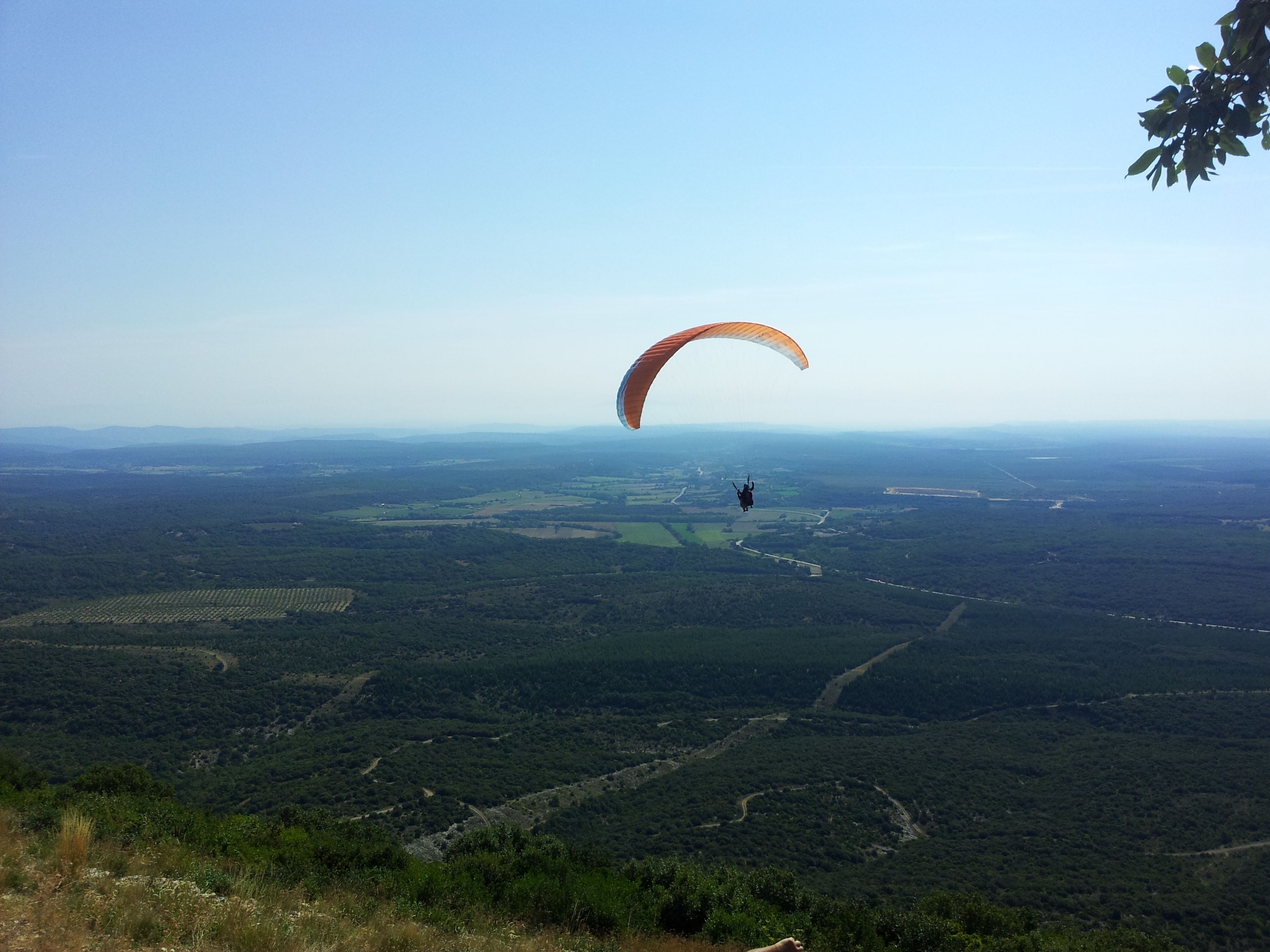 Une personne pratiquant le parapente au-dessus d’un vaste paysage où l’on aperçoit des routes et des champs.
